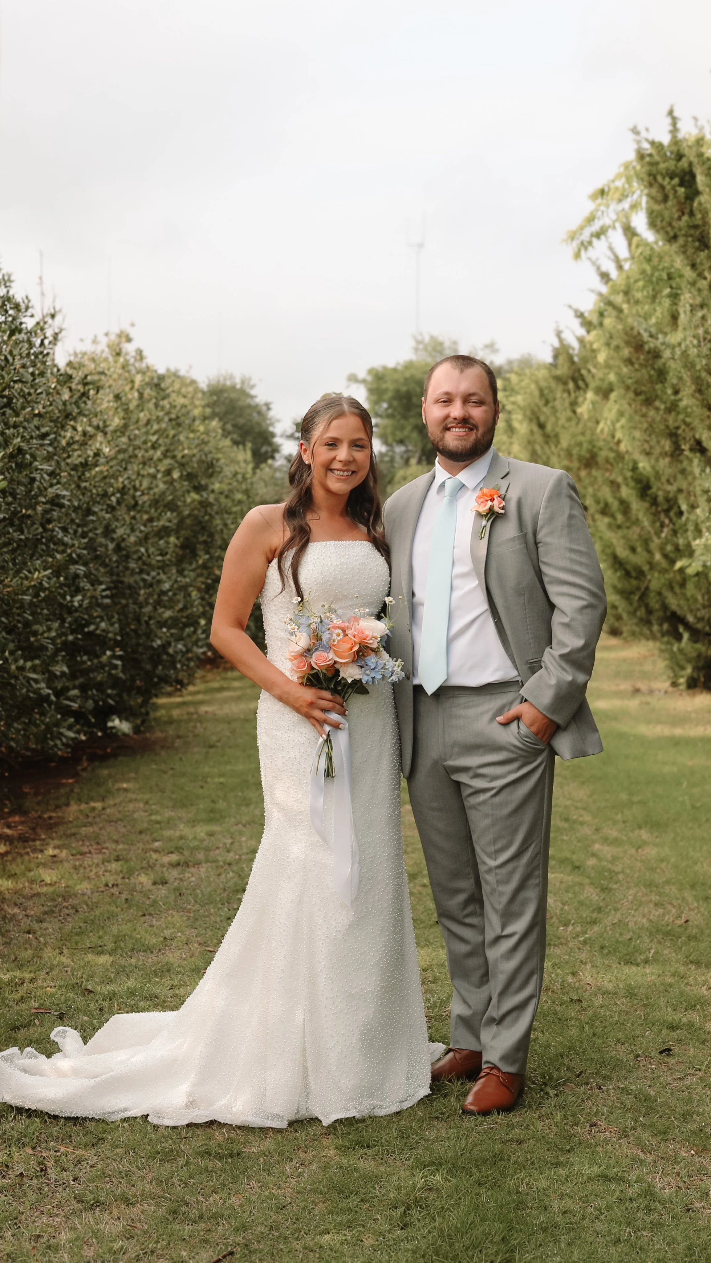 A bride and groom smiling outdoors on their wedding day, standing on grass between trees with cloudy sky in the background. The bride is holding a bouquet of pink, blue, and white flowers, wearing a white strapless wedding gown with a long train. The