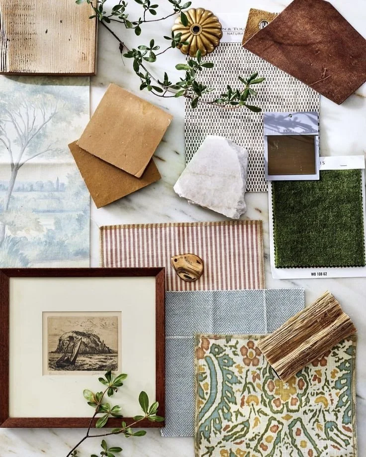 A collection of interior design and decorating samples, including fabric swatches, paint chips, wallpaper, a framed picture, a decorative object, and a plant branch on a white surface.