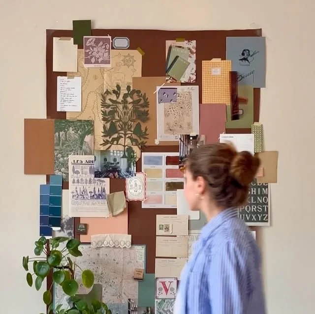 A woman with brown hair tied in a bun looking at a wall decorated with various papers, notes, images, and color swatches, with a potted plant in the foreground.