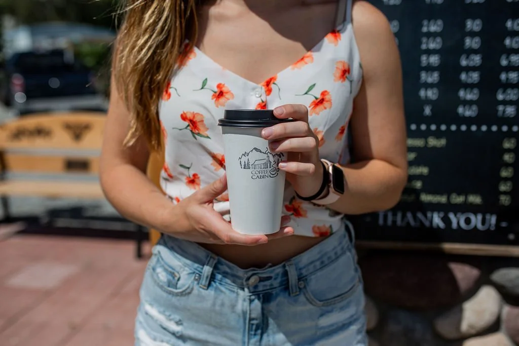 A woman wearing a white tank top with orange floral patterns and denim shorts is holding a take-out coffee cup with a black lid. The background includes a bench and a chalkboard menu with a 'Thank You!' note.