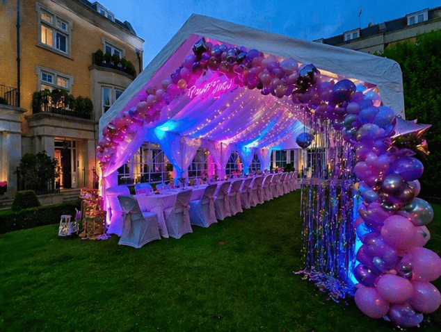 Decorated outdoor event tent with pink and purple balloons, fairy lights, and a long banquet table set for a celebration during dusk.