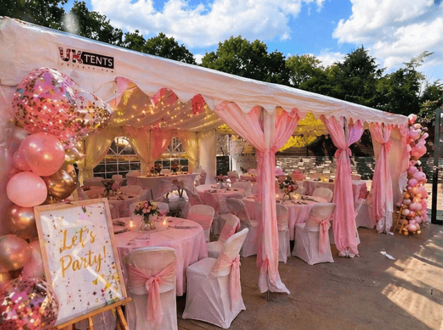 A large outdoor event tent decorated in pink and white with round tables draped in pink tablecloths and chairs covered with white fabric and pink sashes. There are pink, gold, and confetti balloons, floral centerpieces on the tables, and a sign that says "Let's Party!" at the entrance. The tent is set up for a celebration or party, with a partly cloudy sky overhead.