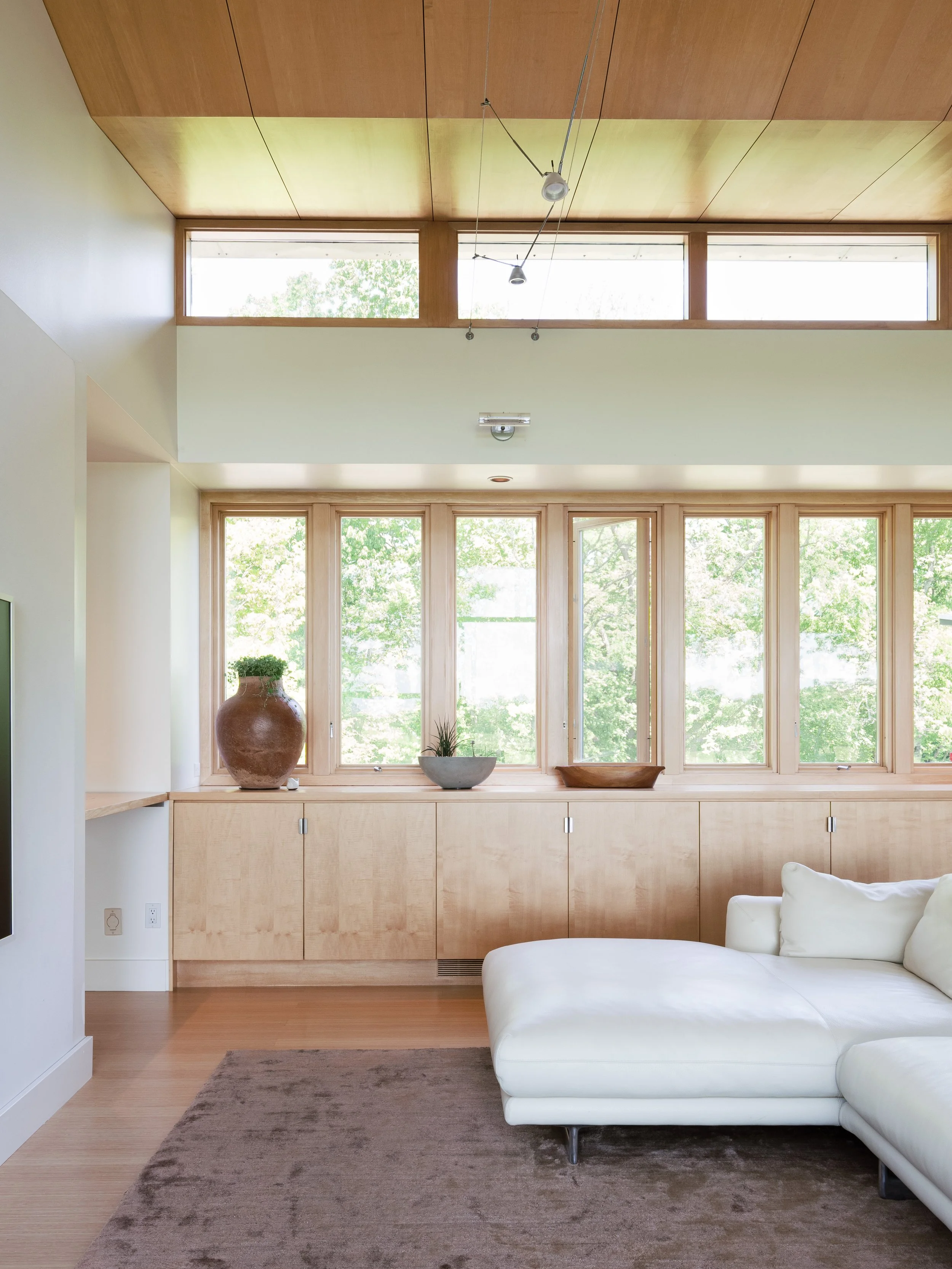 Bright living room with large windows, wooden ceiling, white sectional sofa, and minimal decor including vases and bowls.