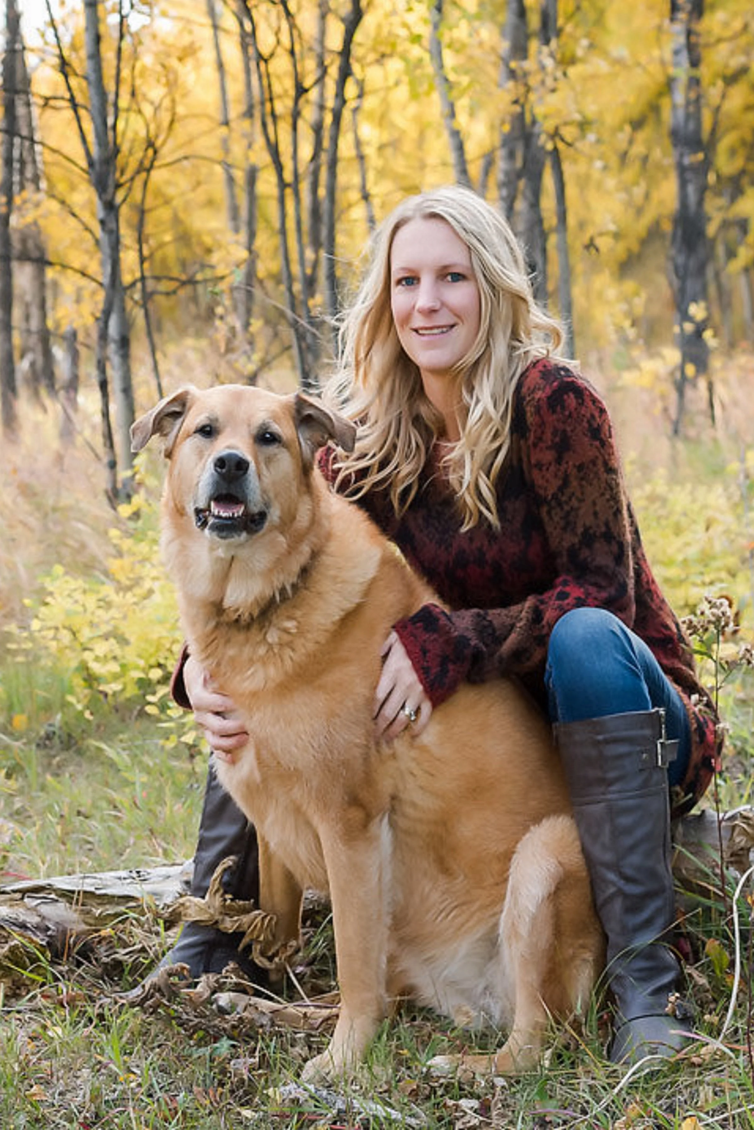A woman kneels next to a large tan dog in an autumn forest with yellow and orange leaves.