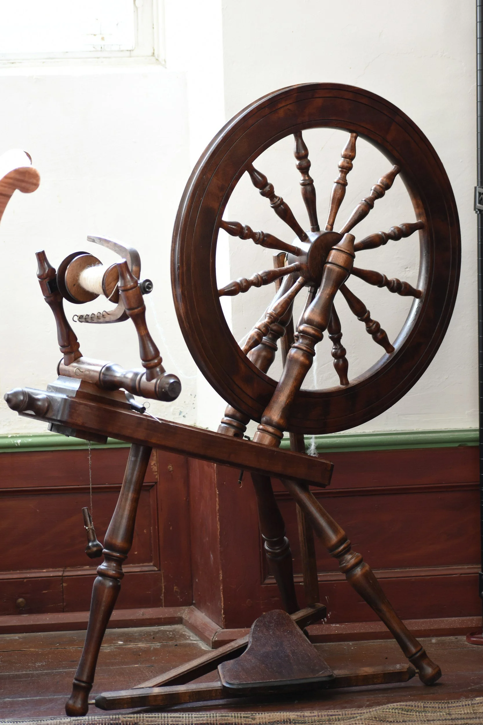 An antique spinning wheel with a large wooden wheel, an adjustable tension arm, and a foot pedal, set against a white wall near a window.