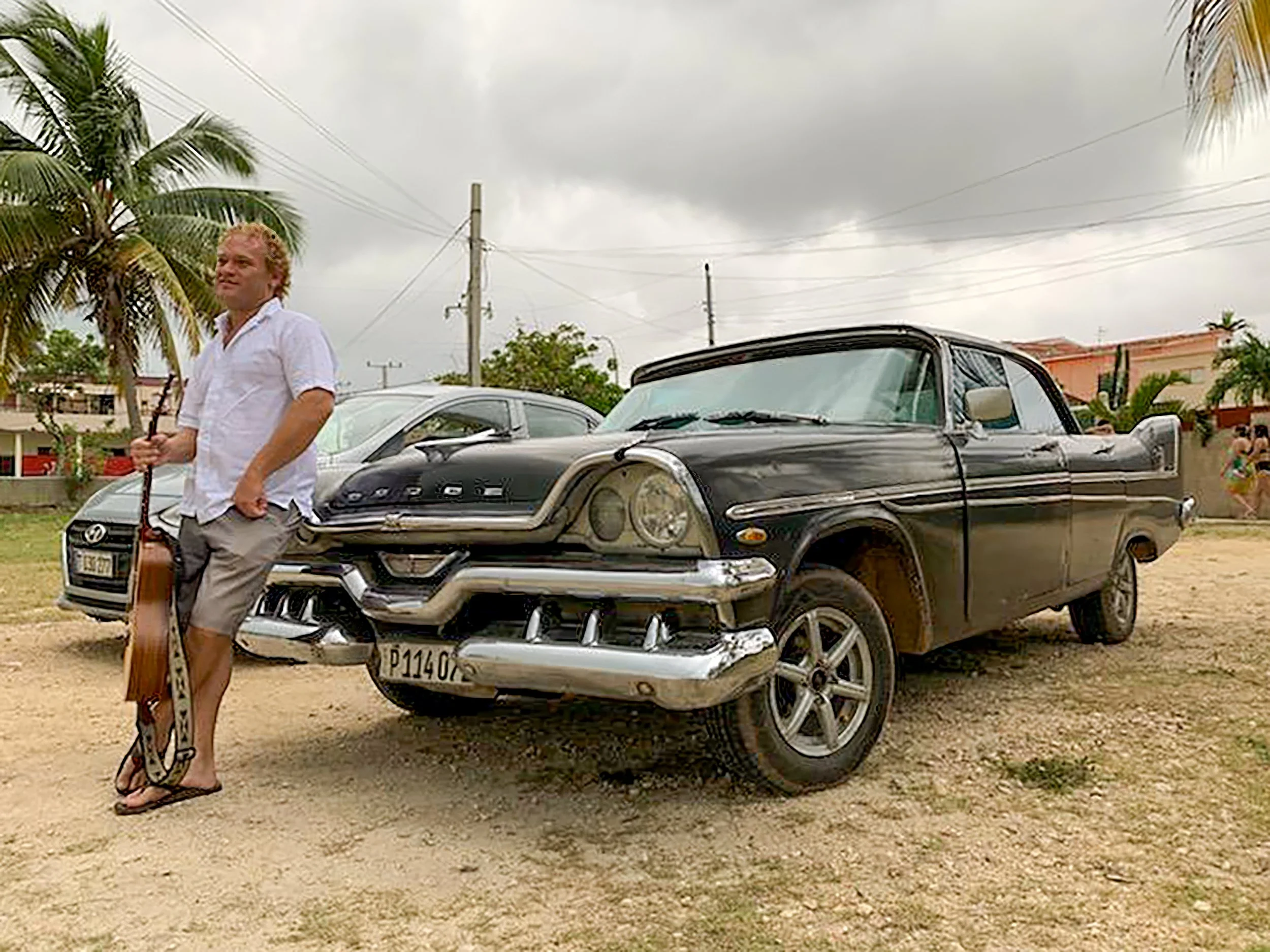 A man standing next to a vintage black car holding a guitar and a woman walking in the background with other cars and palm trees.