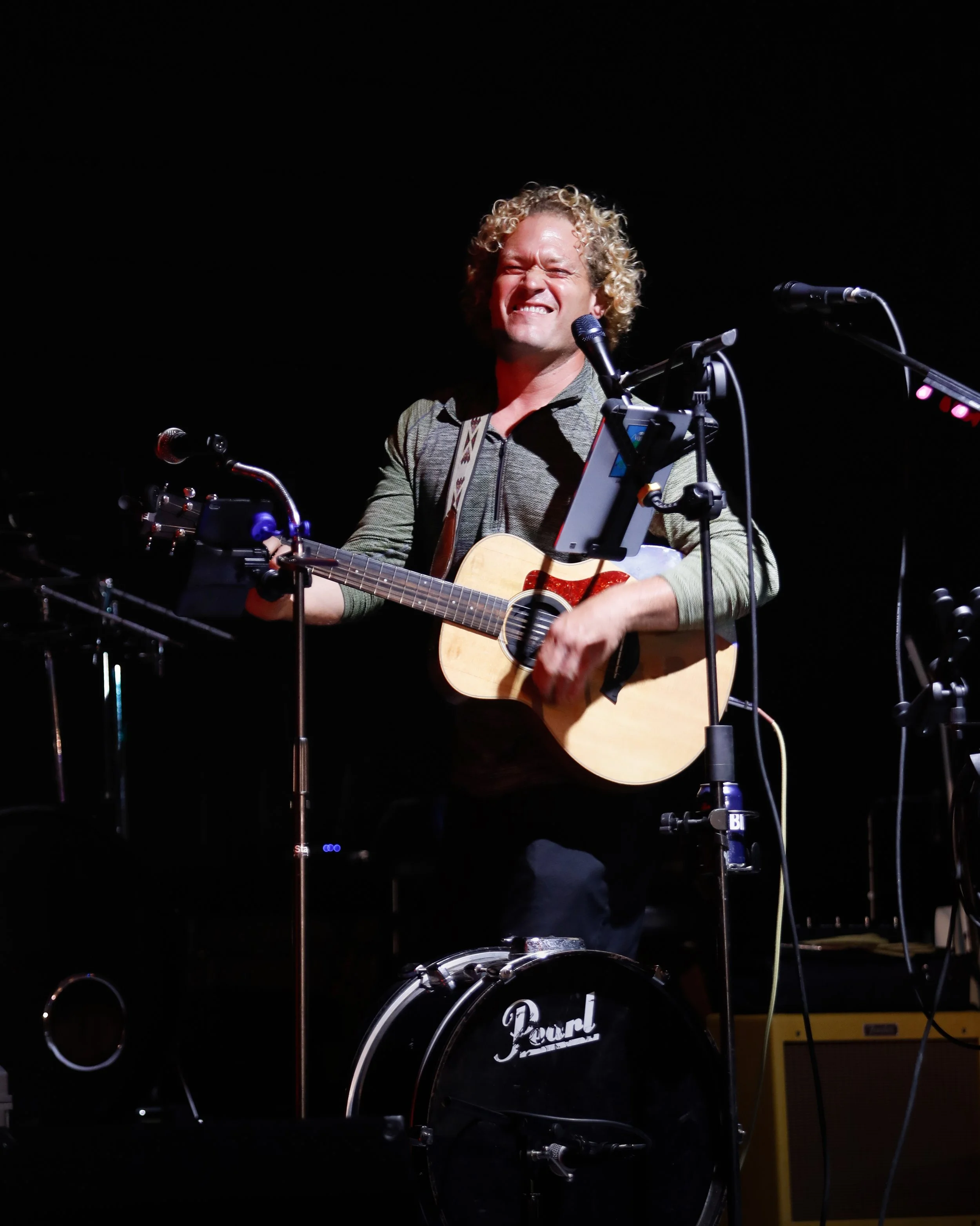 A man with curly blond hair playing an acoustic guitar on stage, smiling with eyes closed, surrounded by microphone stands and musical equipment.