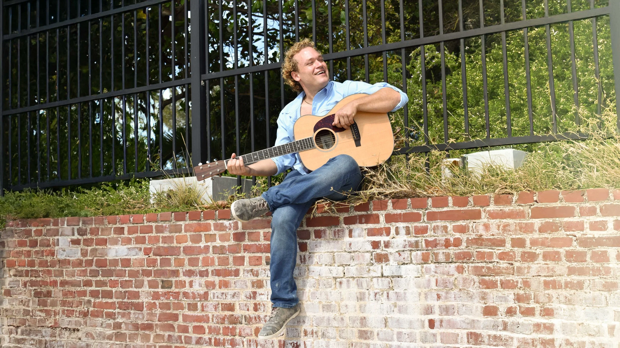 A man sitting on a brick wall, playing an acoustic guitar with a broad smile.