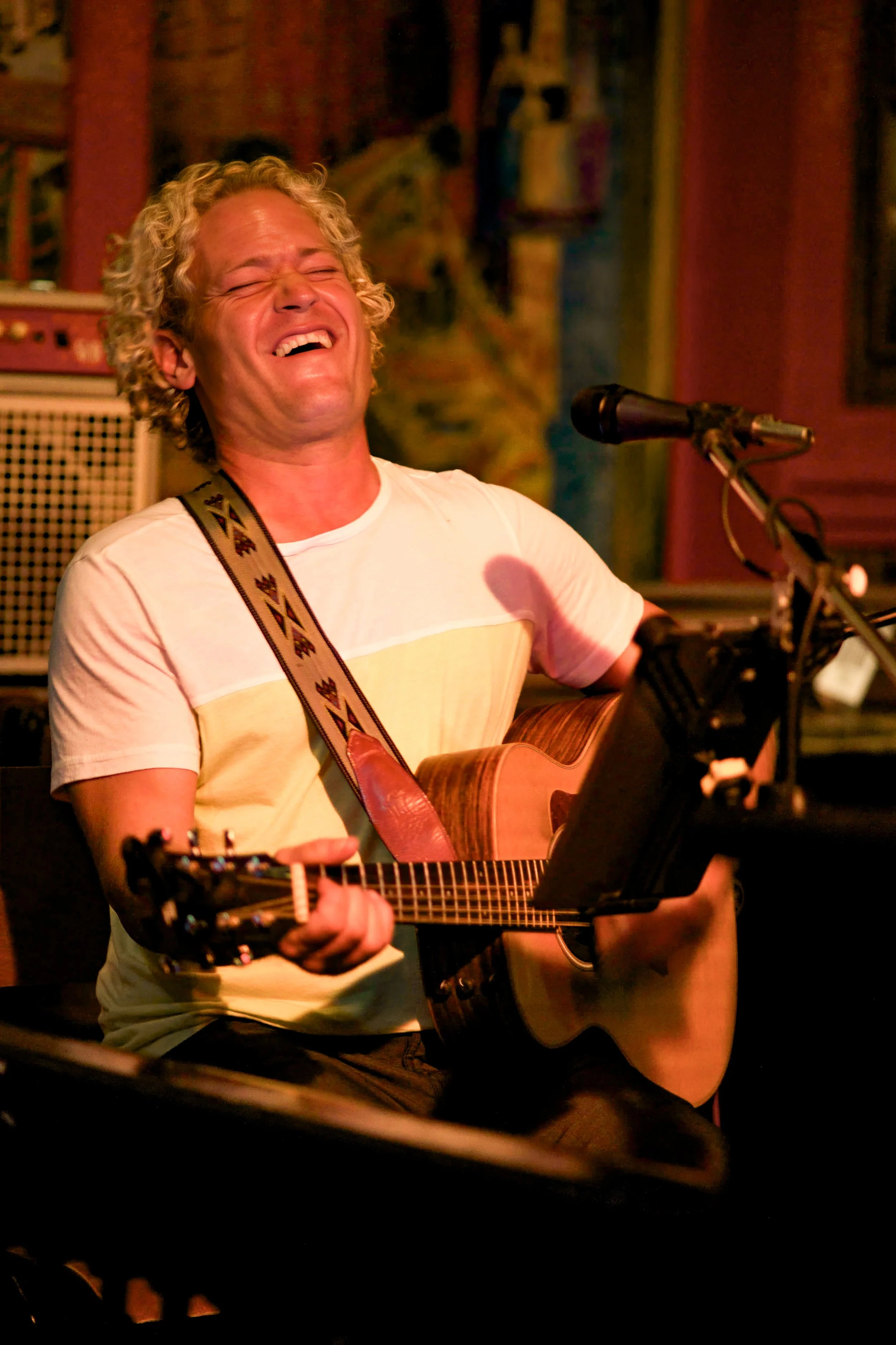 A man with curly blonde hair passionately singing and playing an acoustic guitar during a live music performance in a dimly lit venue.