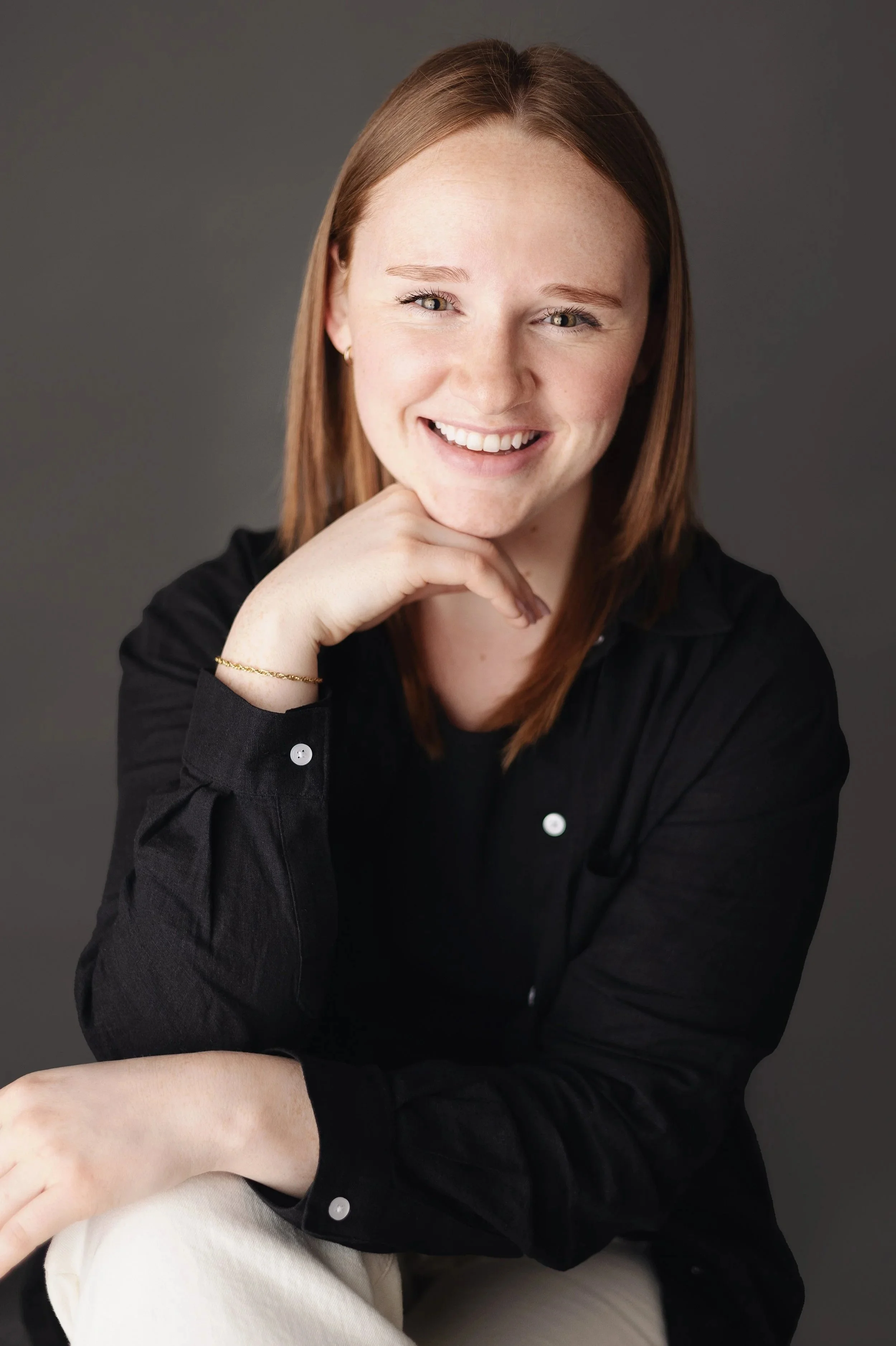 Portrait of a woman with red hair, smiling, wearing a black shirt, posing with her hand under her chin against a dark background.