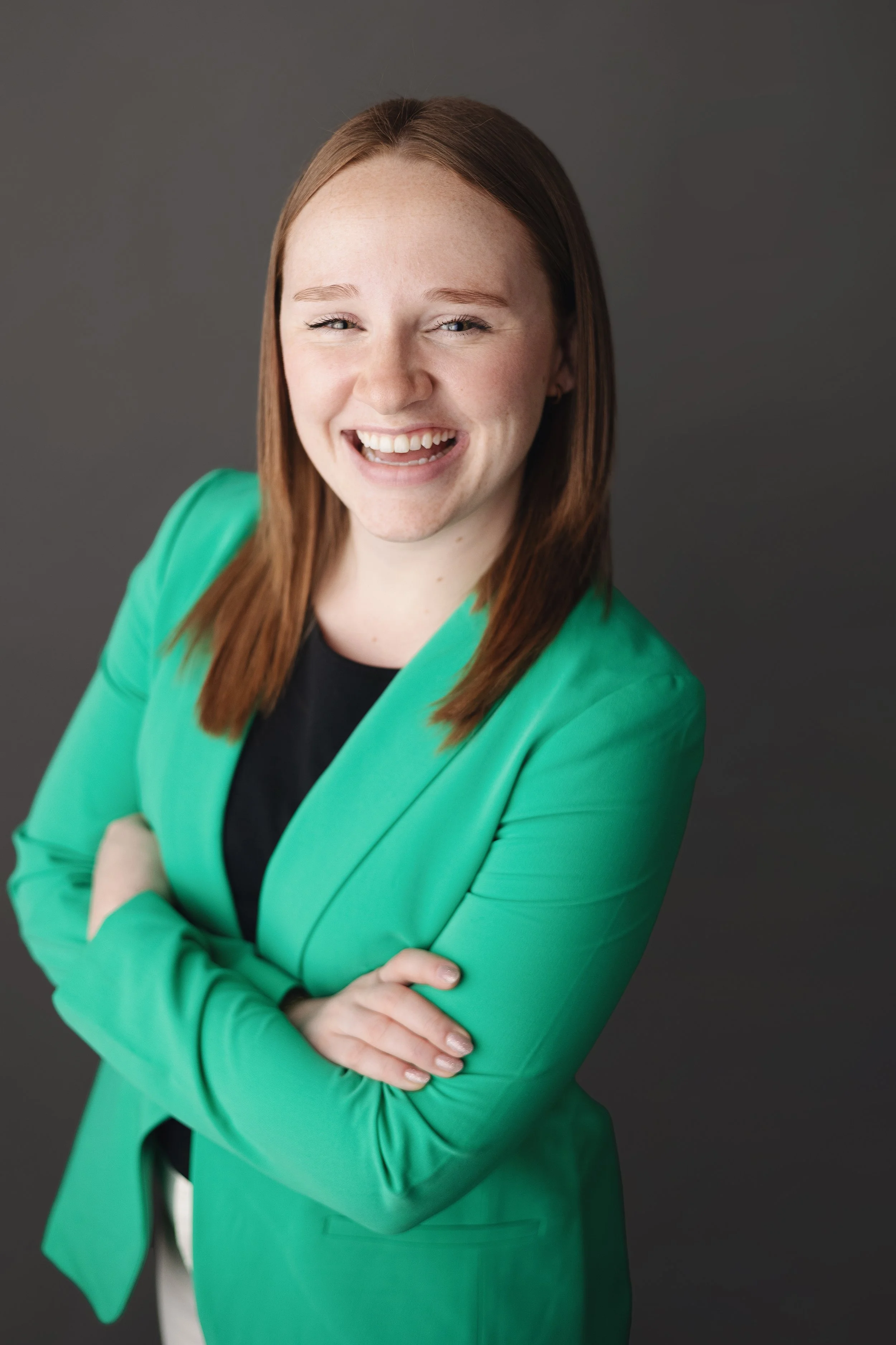 A woman with red hair wearing a green blazer and black top, smiling with arms crossed, in front of a dark grey background.