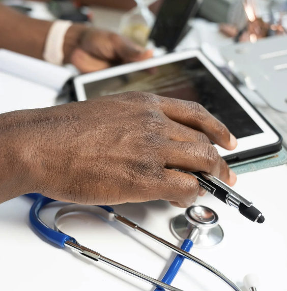 Close-up of a person's hand resting on a tablet with a stylus, with a stethoscope nearby, on a white table.