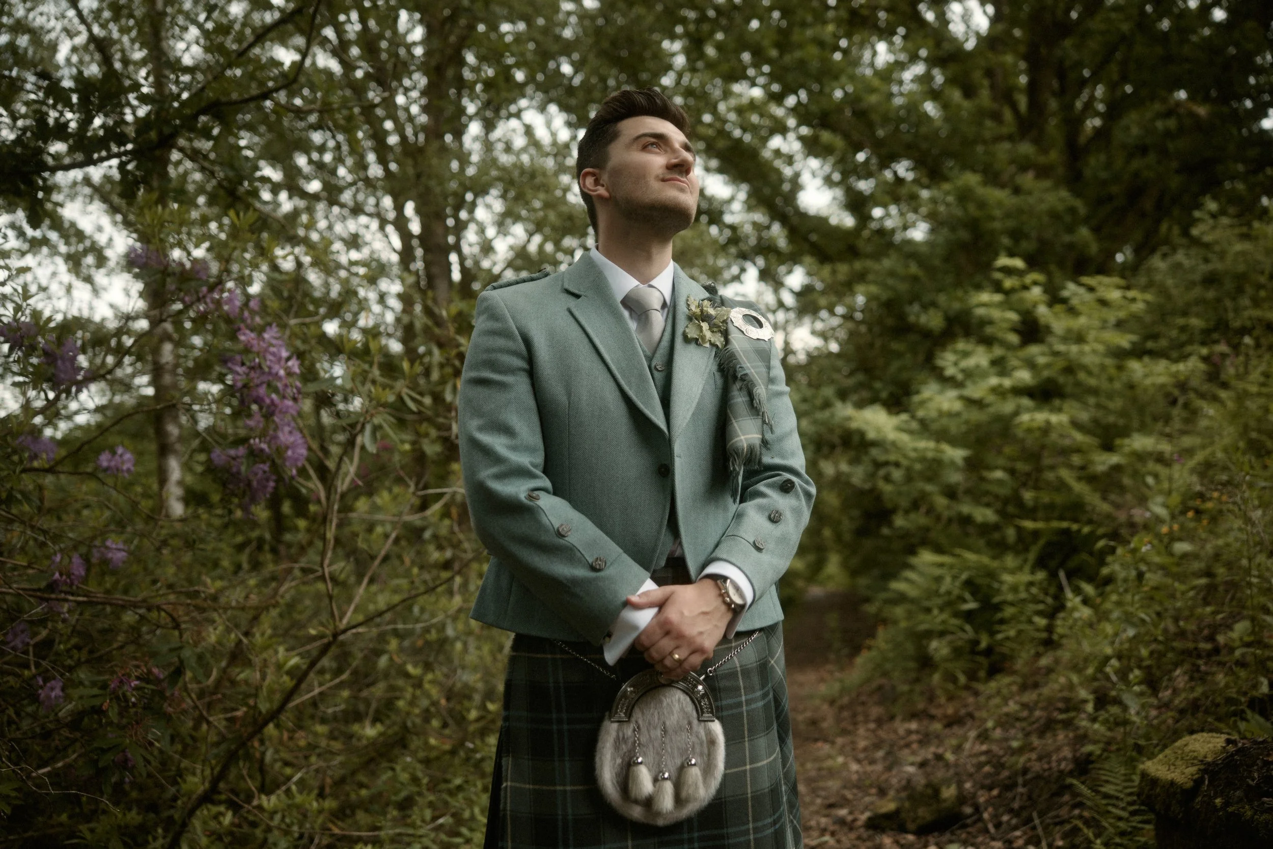 A man wearing traditional Scottish attire, including a kilt and jacket, standing in a wooded outdoor setting with trees and greenery, looking upward.