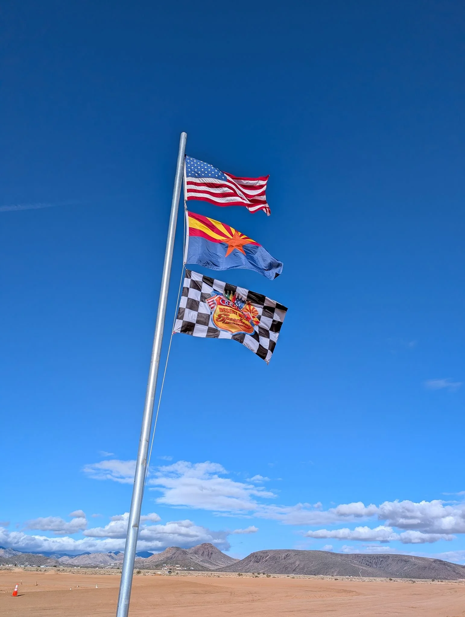 Three flags on a tall flagpole against a blue sky with mountains in the background; the top flag is the United States flag, the middle is a flag with a red sunburst on a blue and yellow background, and the bottom is a checkered flag with a logo.