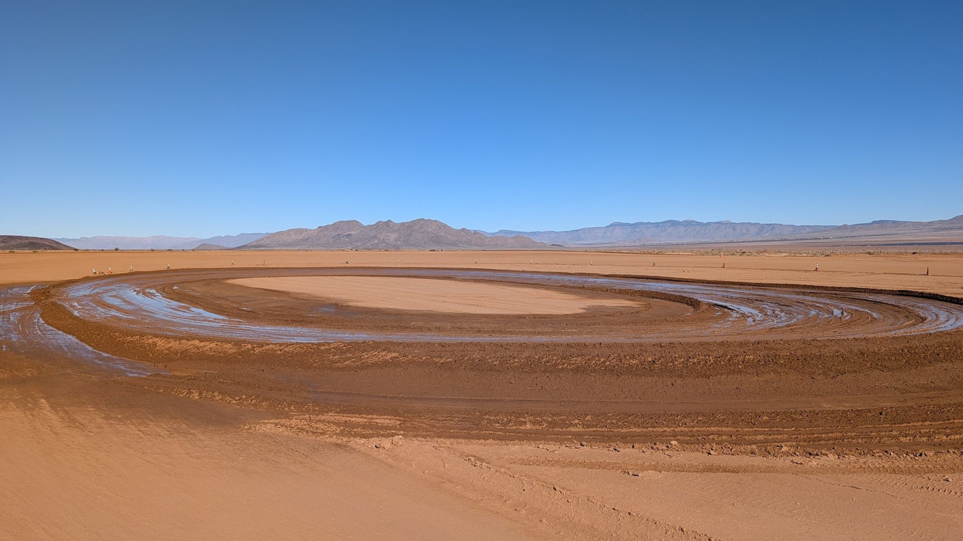 A desert landscape with a dirt racetrack, mountains in the distance, and a clear blue sky.