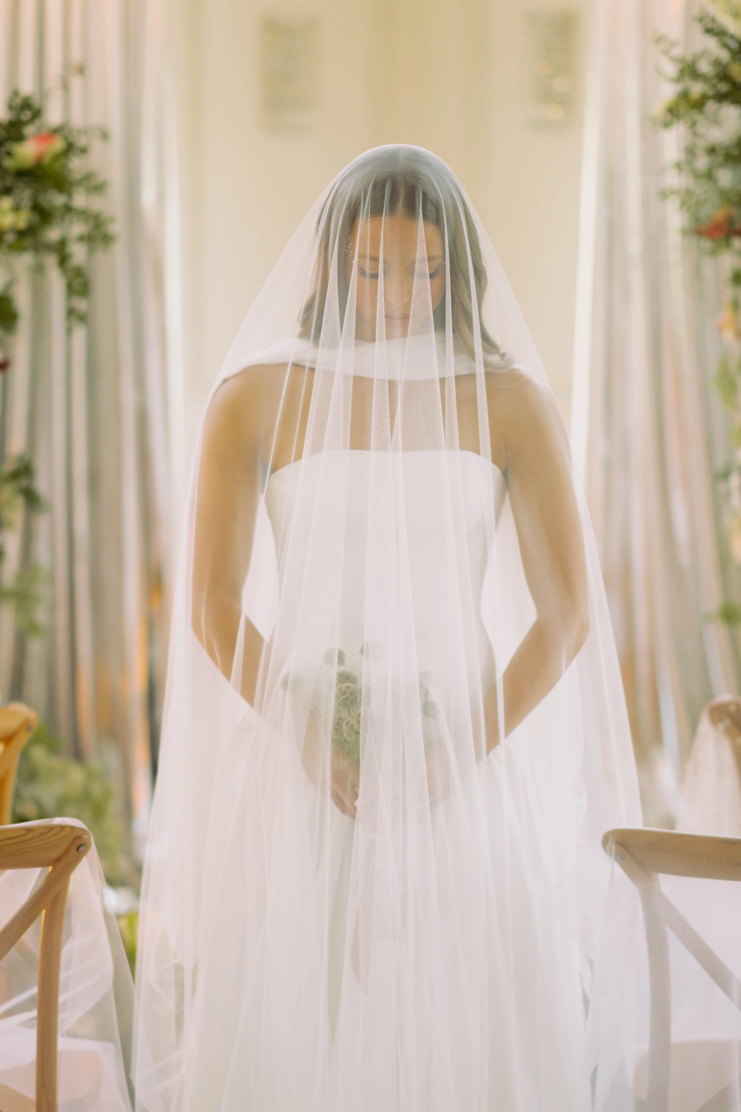 A woman in a wedding dress with a veil standing inside a softly lit room, surrounded by curtain panels and greenery.
