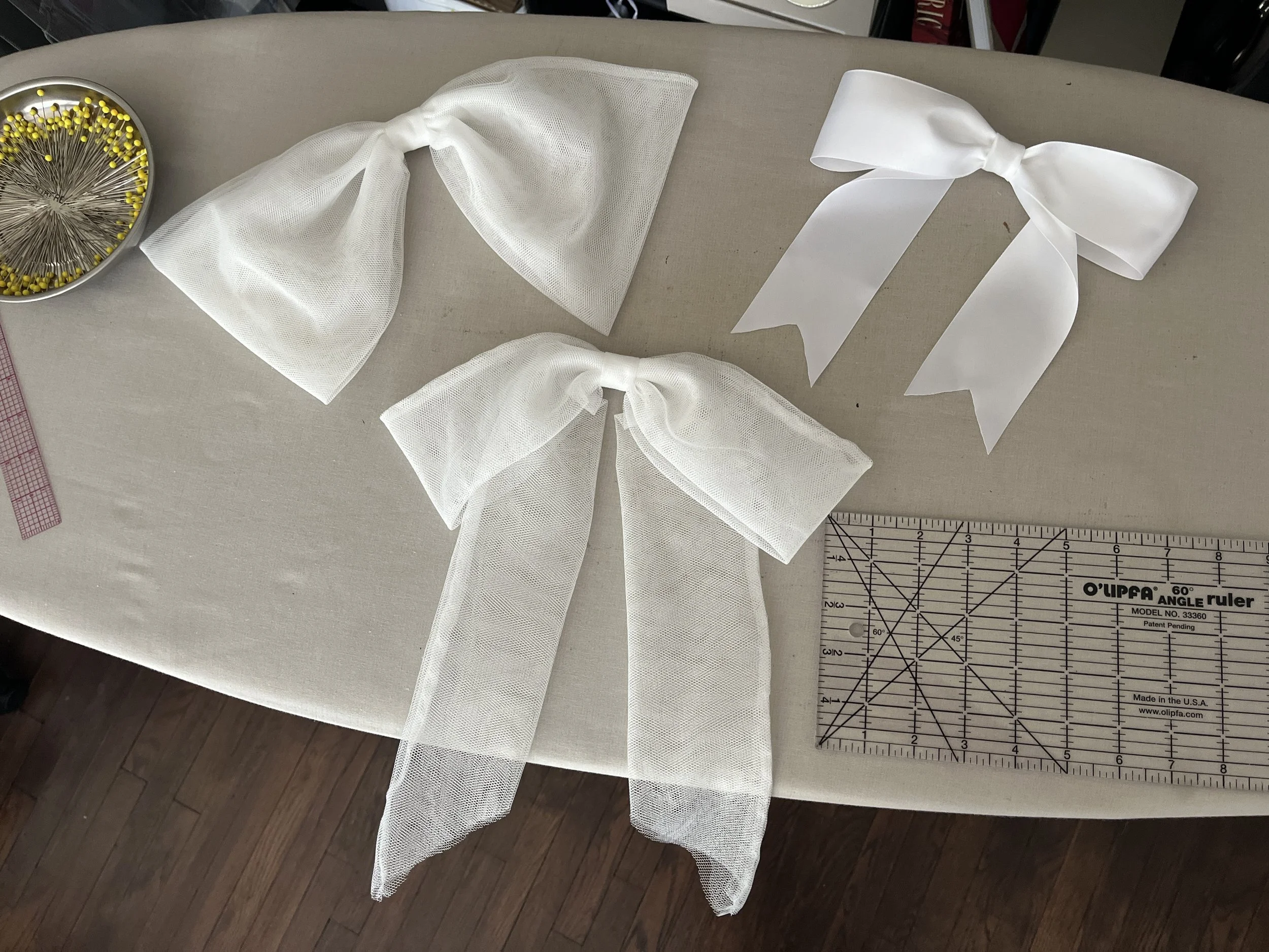 A work table with sewing supplies including three white bows made of net fabric, a bowl of yellow-headed pins, and a clear quilting ruler.