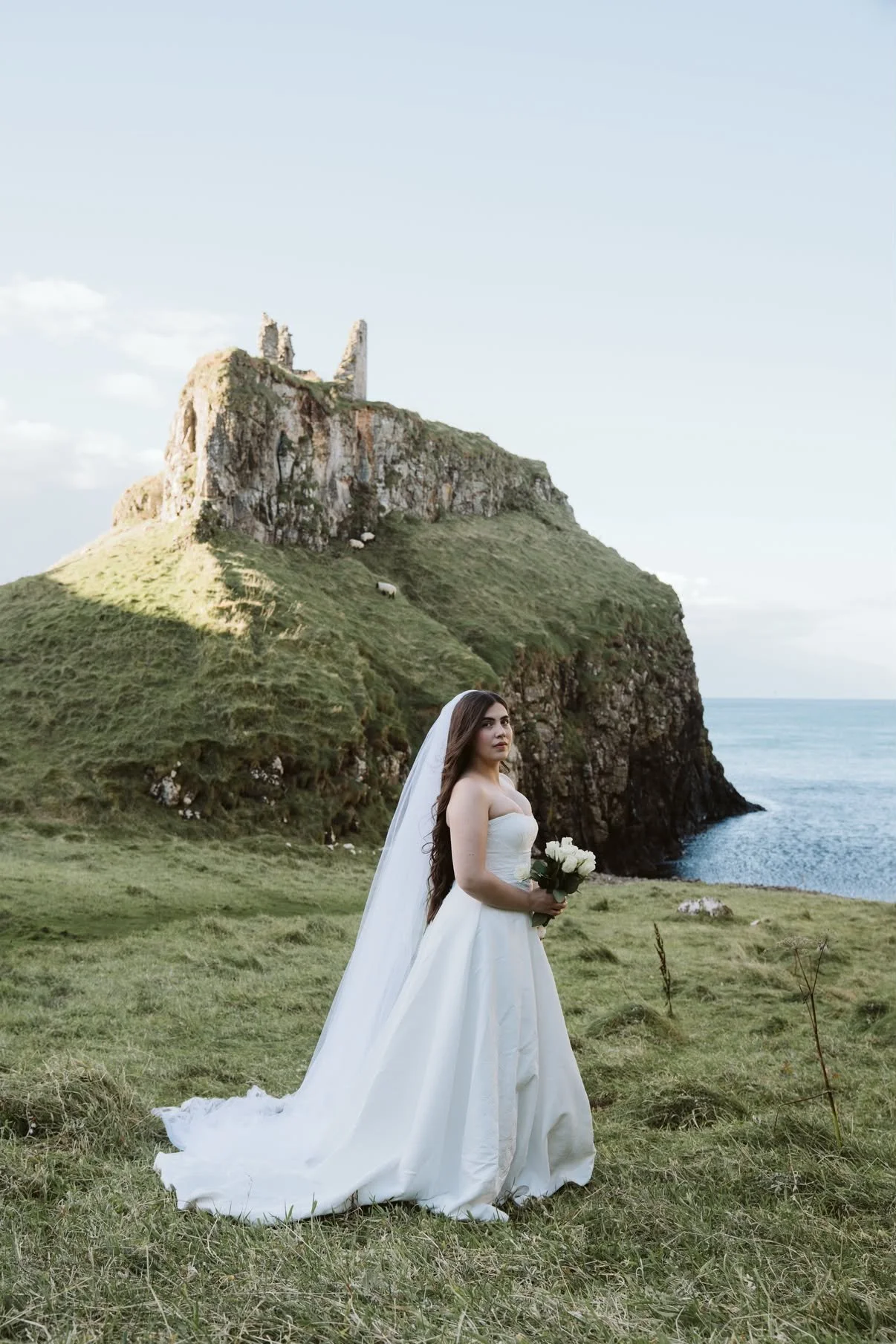 A young woman in a white wedding dress and long veil holding a bouquet of white roses stands on a grassy field, with green hills and a castle ruin on a hill in the background, overlooking cliffs down to the ocean