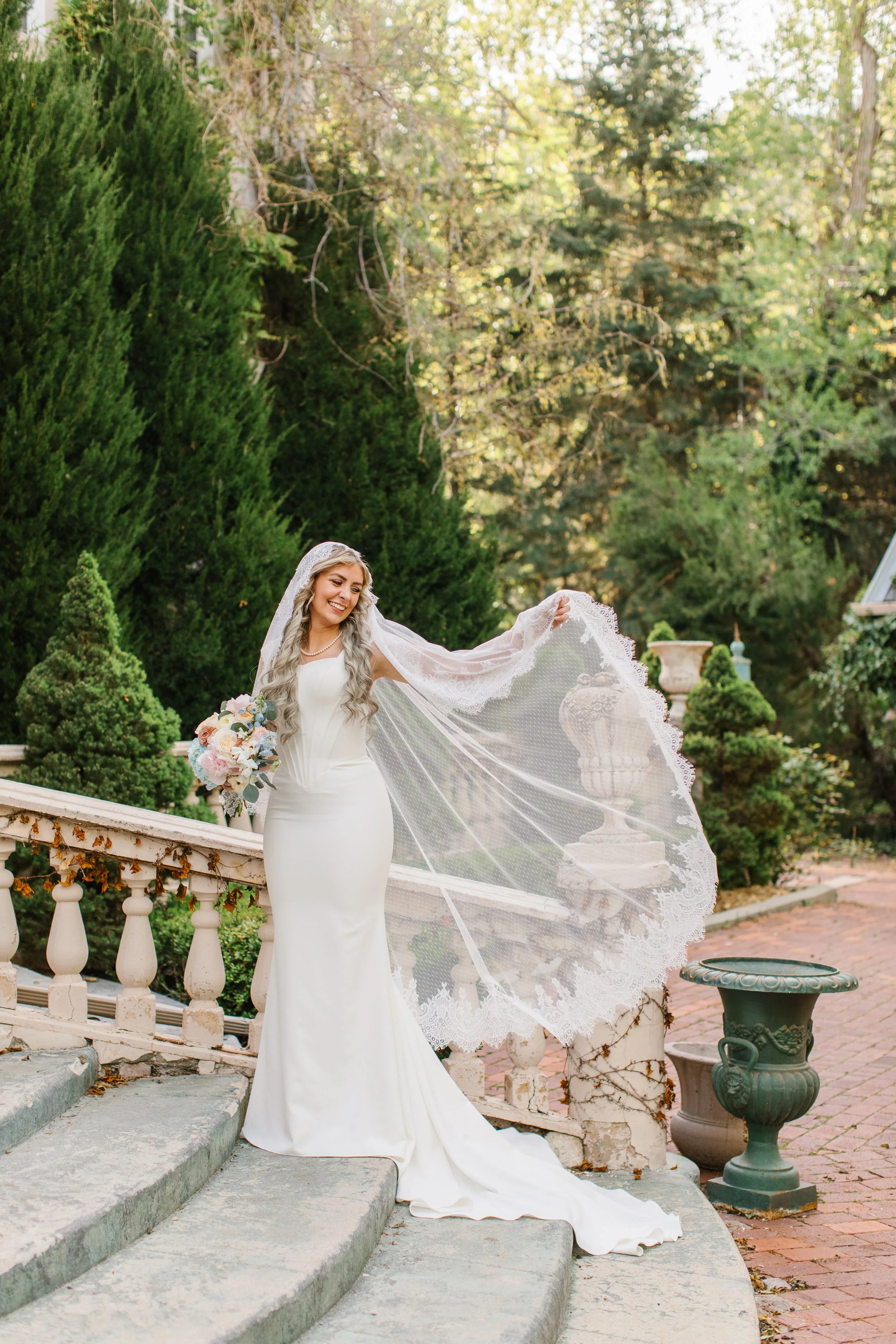 A young woman in a wedding gown and lace trimmed mantilla veil stands on a set of outdoor stone steps. She holds the veil out to her side so it sweeps behind and to the side of her
