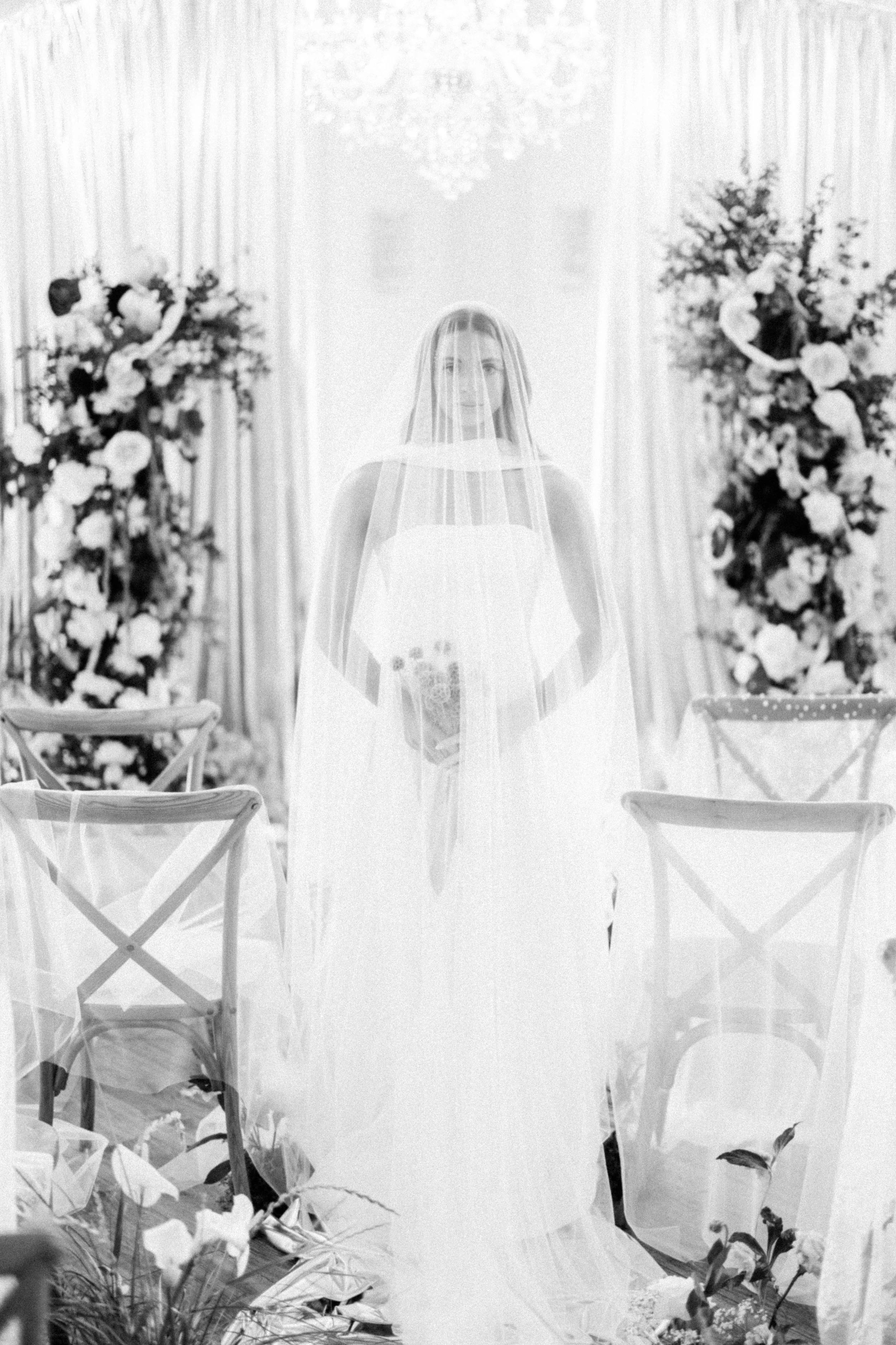A woman in a wedding gown with a veil over her head and the front of her dress, in front of a decorated altar with floral arrangements and chairs.
