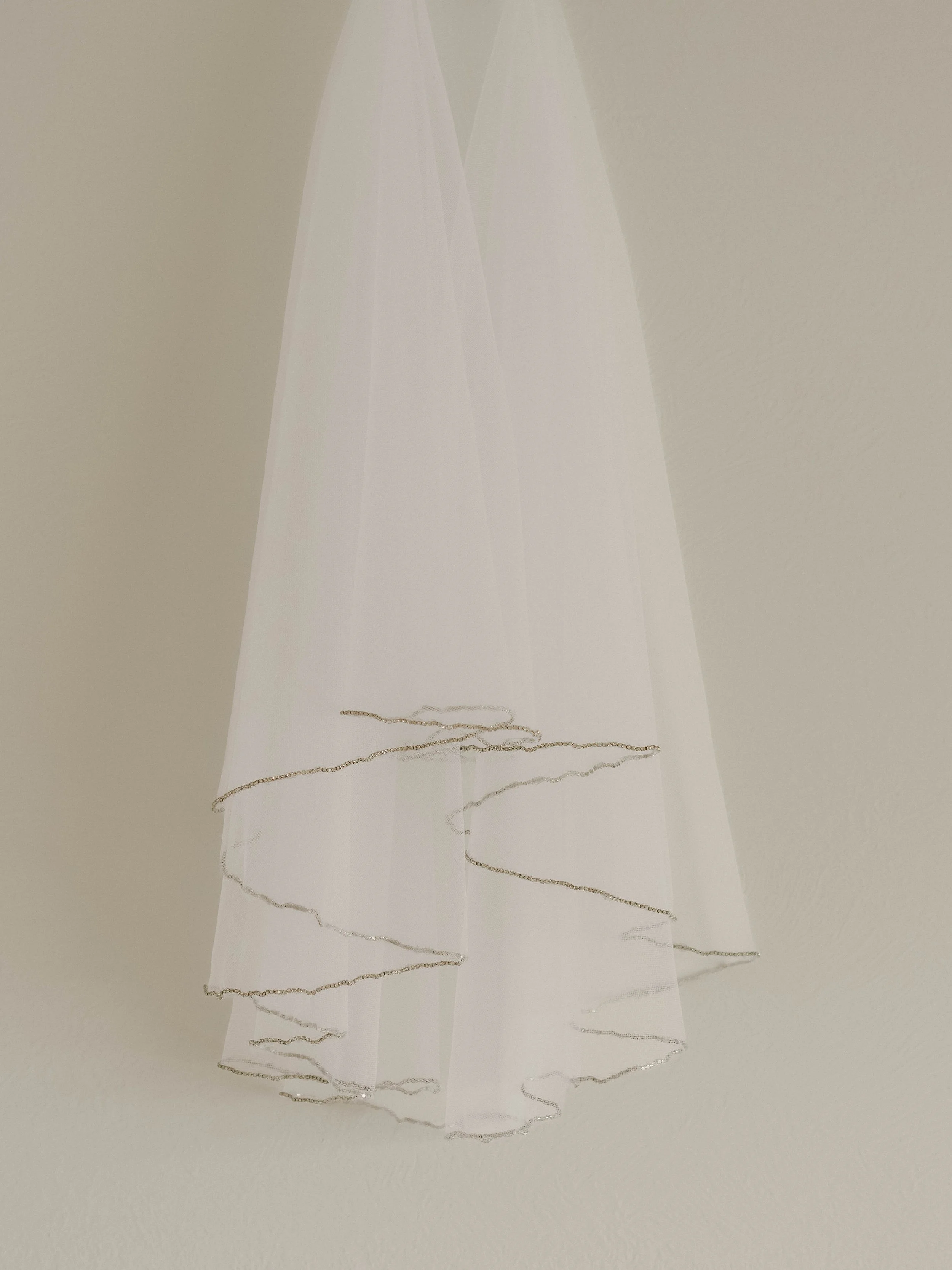 A white veil with delicate silver embroidery, hanging in front of a plain wall.
