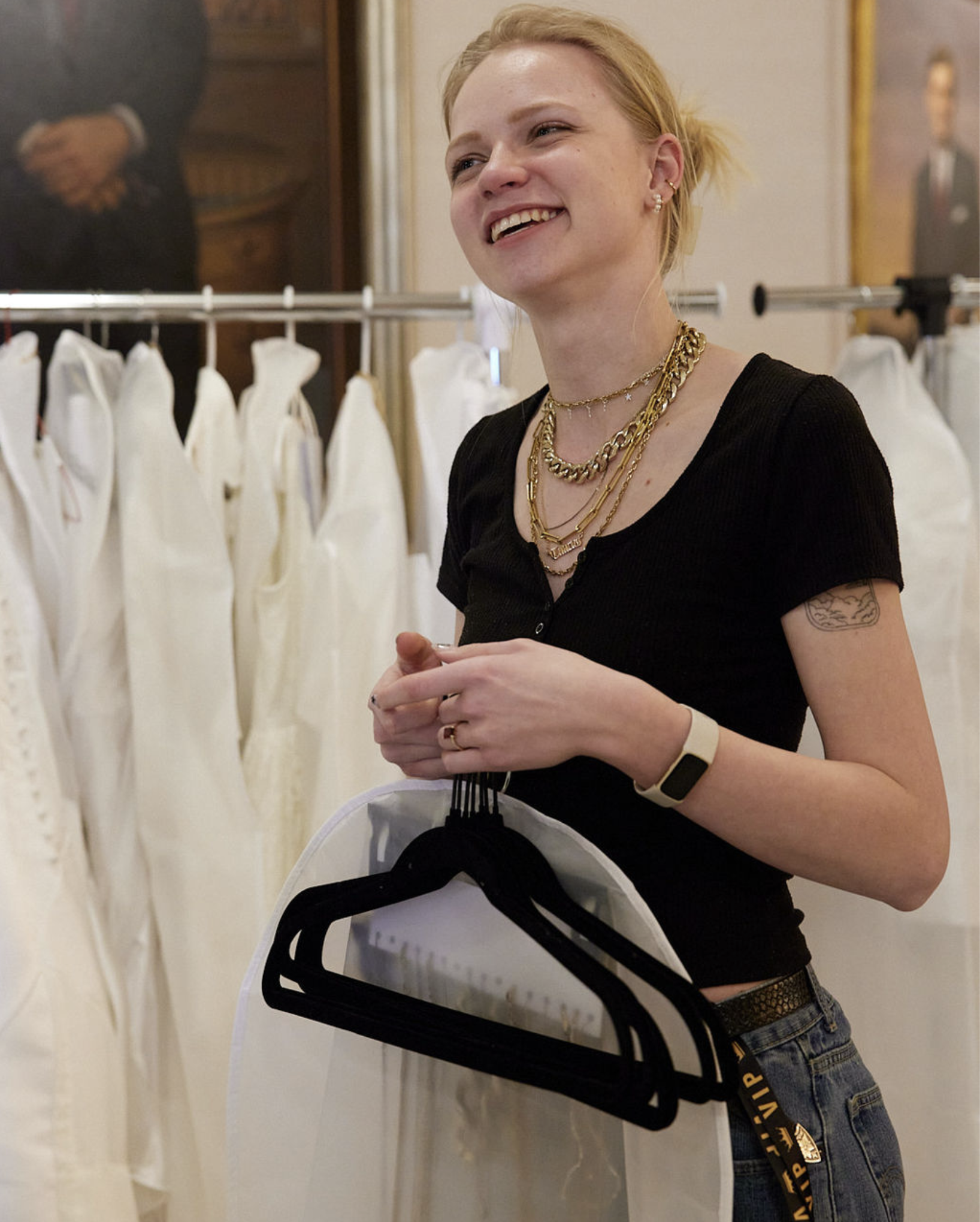 Young woman with blonde hair and tattoos smiling while holding clothing and hangers in front of a clothing rack