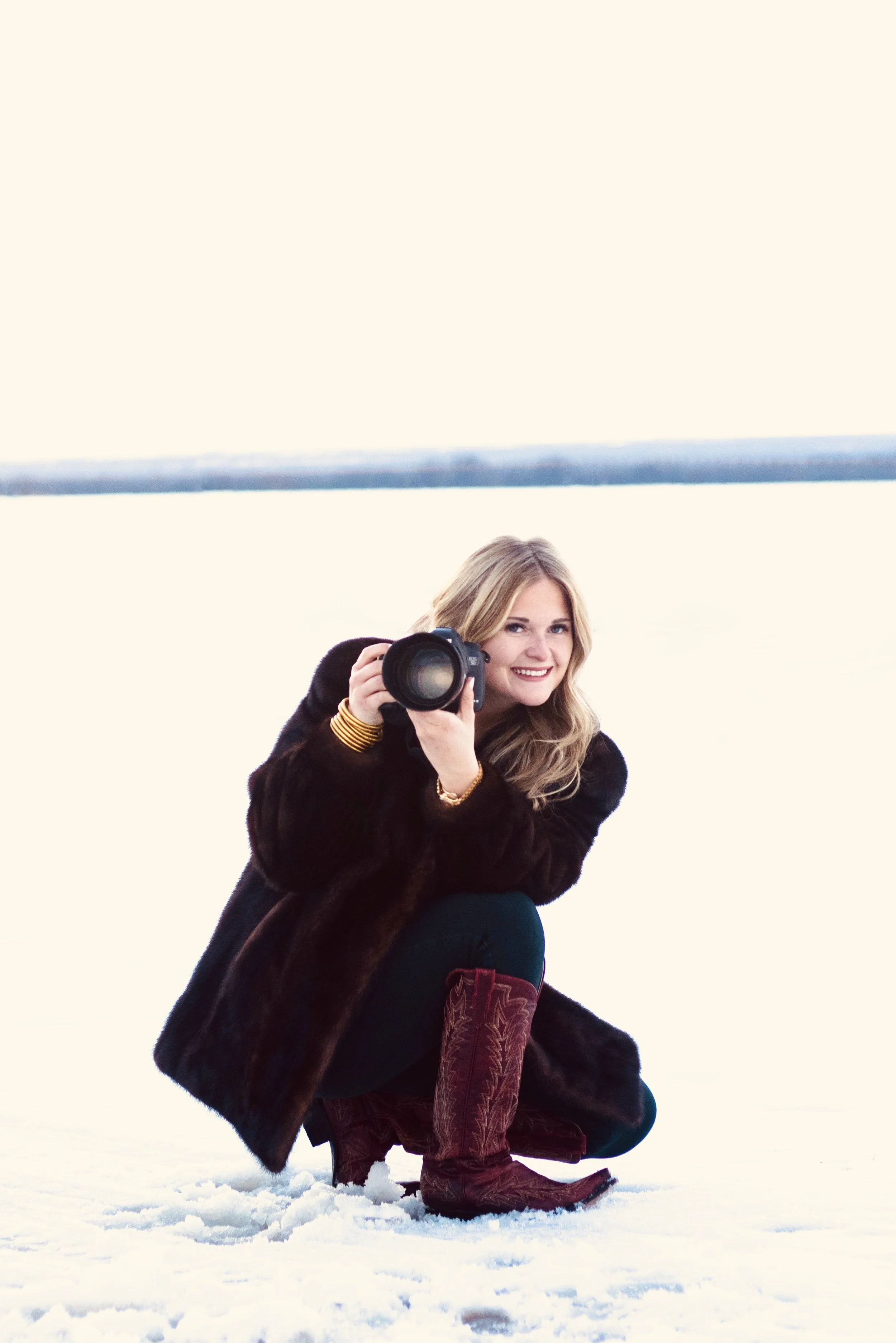 A woman crouching on snow, holding a camera and smiling outdoors.