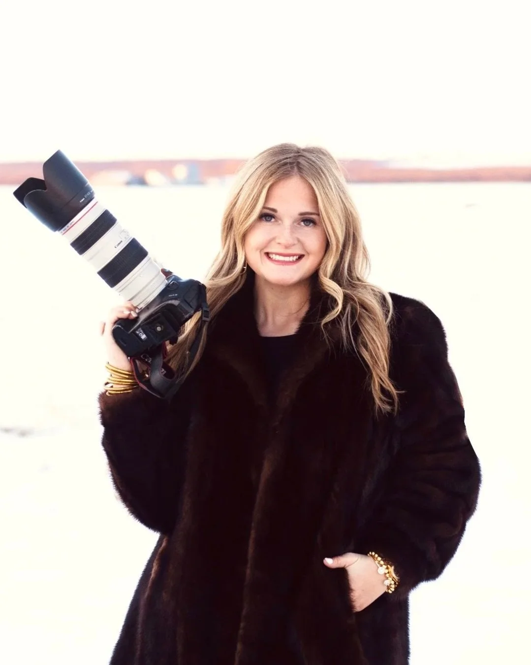 A woman smiling outdoors, holding a professional camera with a large telephoto lens on her shoulder, dressed in a dark fur coat.