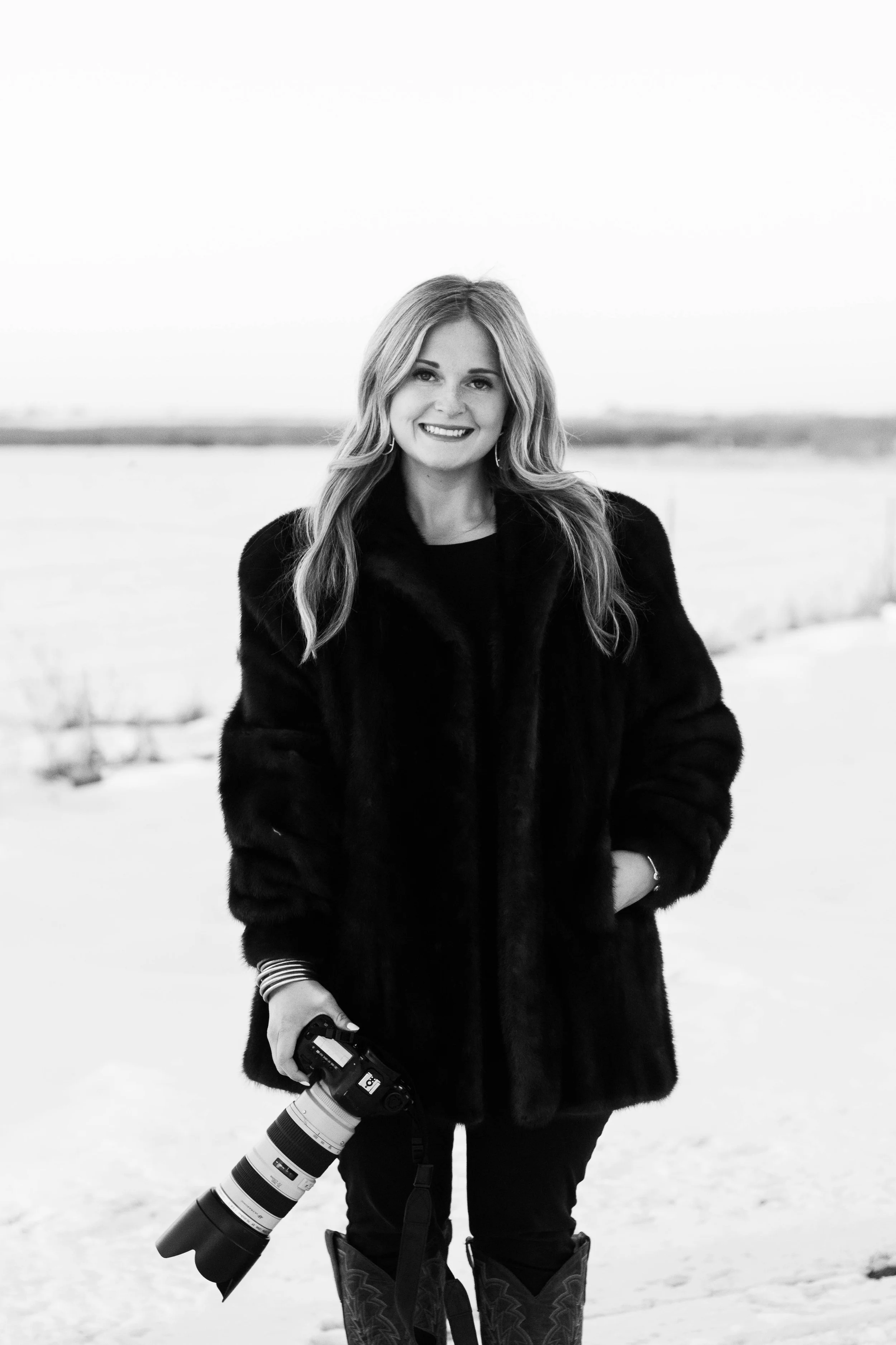 A smiling woman stands outdoors in a snowy landscape, wearing a dark fur coat and holding a professional camera with a telephoto lens.