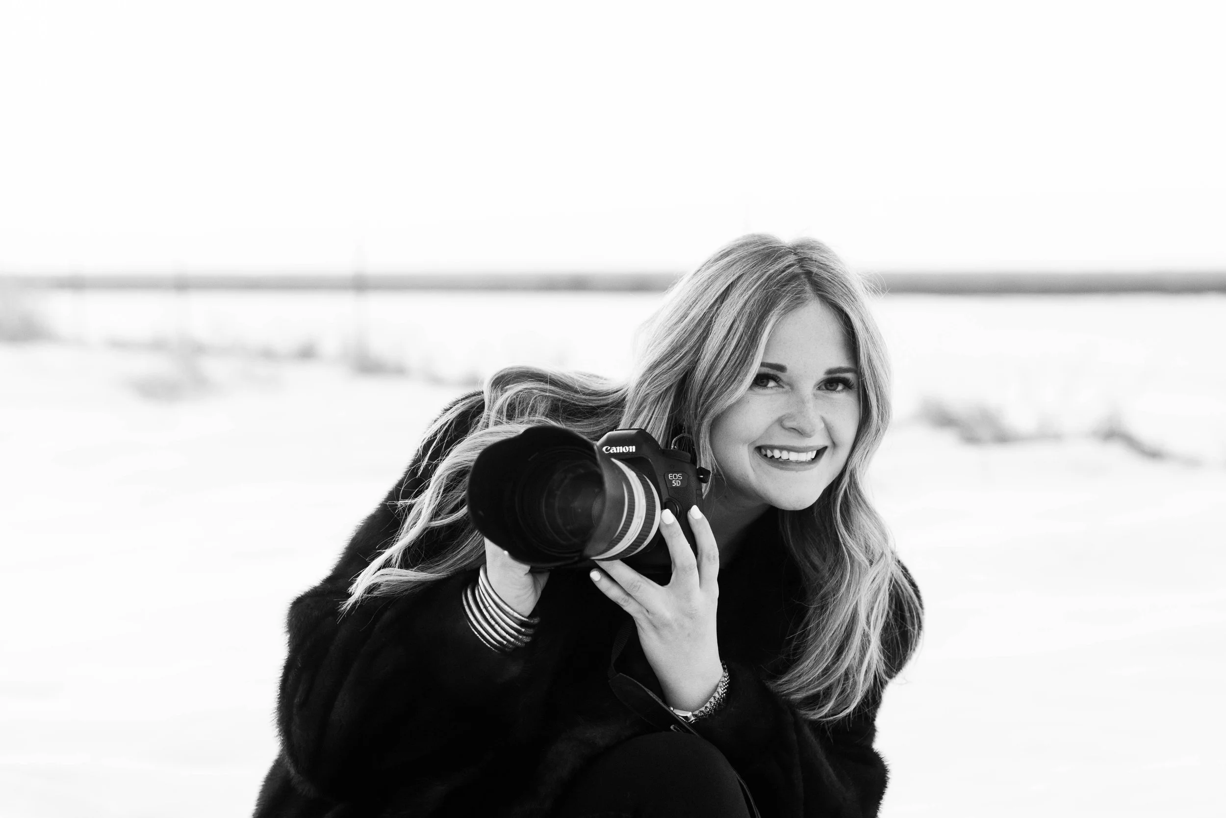 A woman smiling and holding a Canon EOS 5D camera outdoors in a black and white photo.