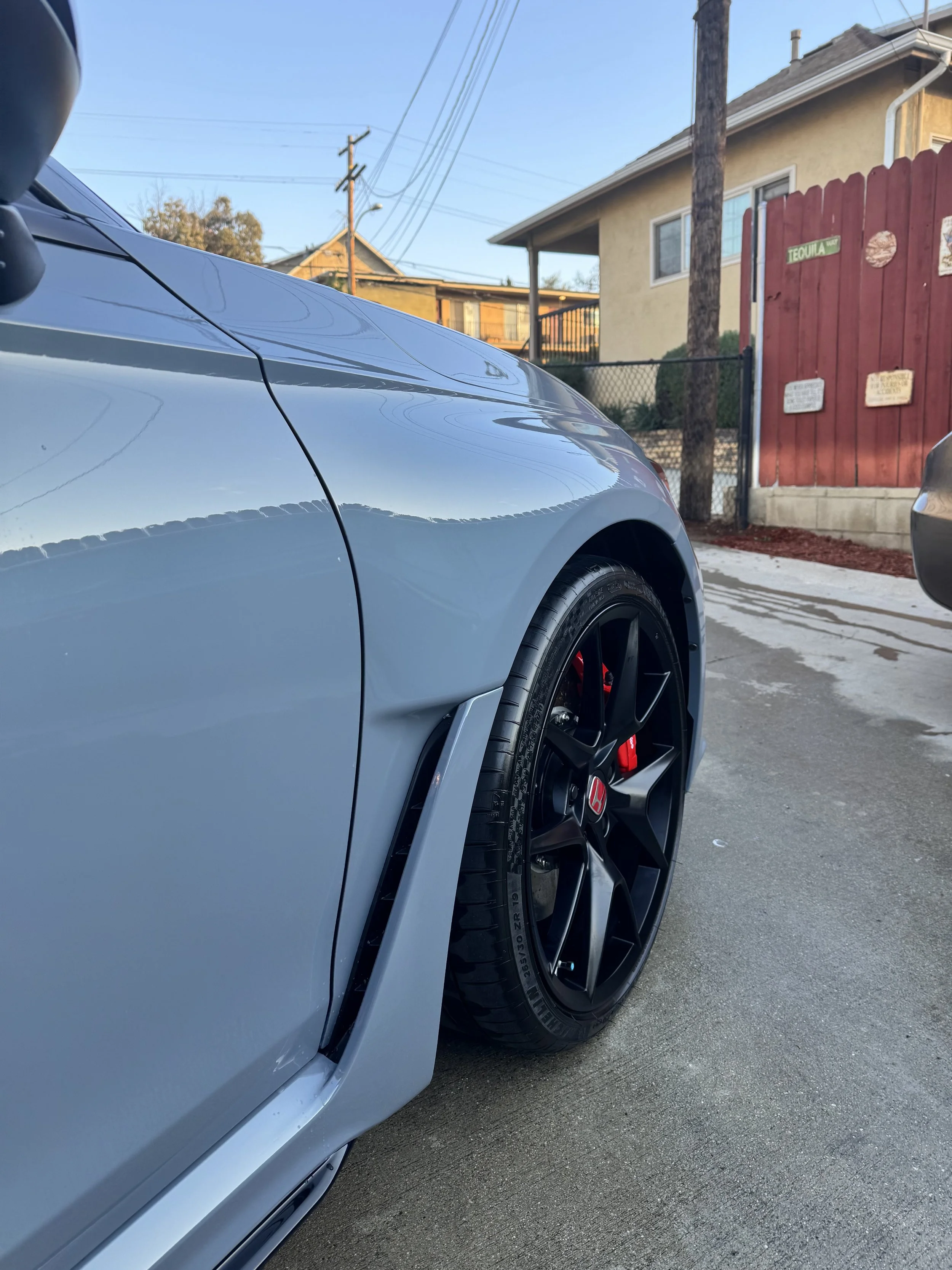 Close-up of a gray sports car with black wheels and red brake calipers parked on a concrete driveway with a house and a red fence in the background.