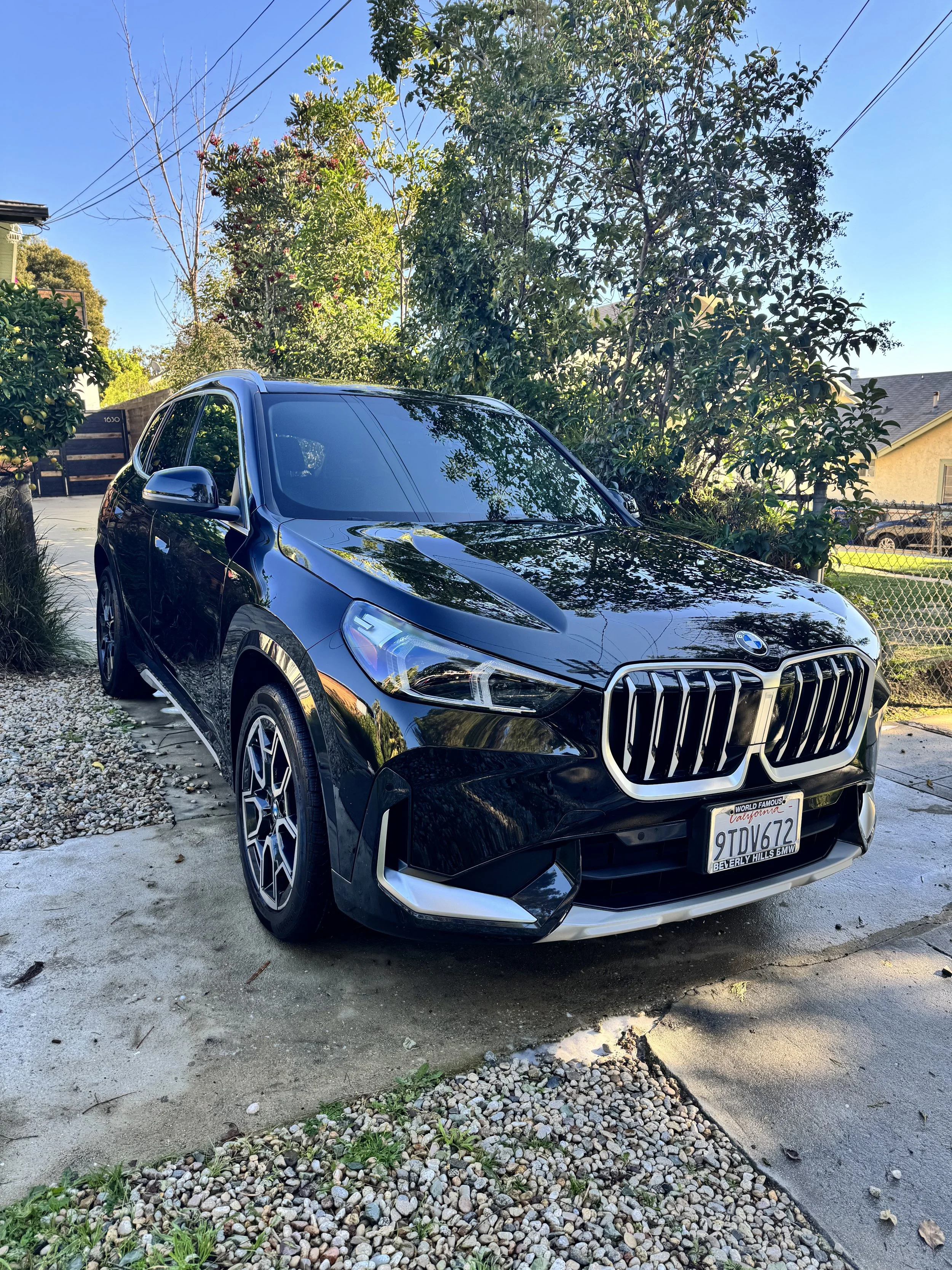 A black BMW SUV parked on a driveway with trees and houses in the background under a clear blue sky.