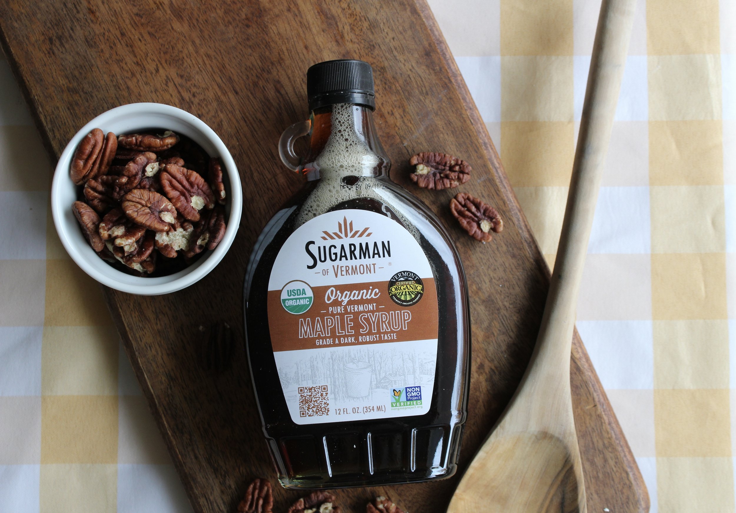 A bottle of Sugarman Vermont organic maple syrup on a wooden surface, with a small bowl of pecans and a wooden spoon nearby, and a checkered tablecloth underneath.