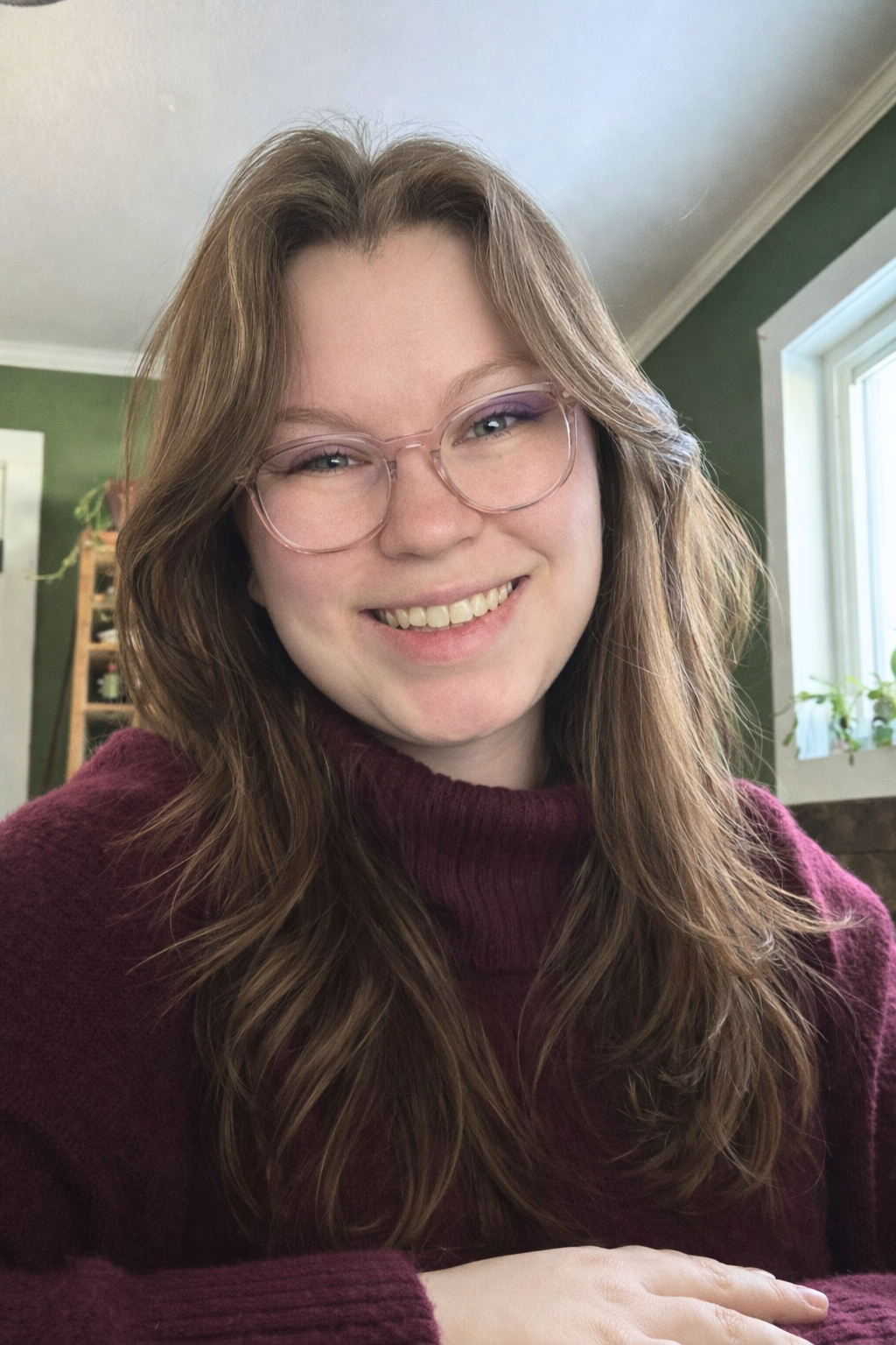 Smiling young woman with glasses and wavy brown hair, wearing a maroon sweater, indoors with green walls and a window.