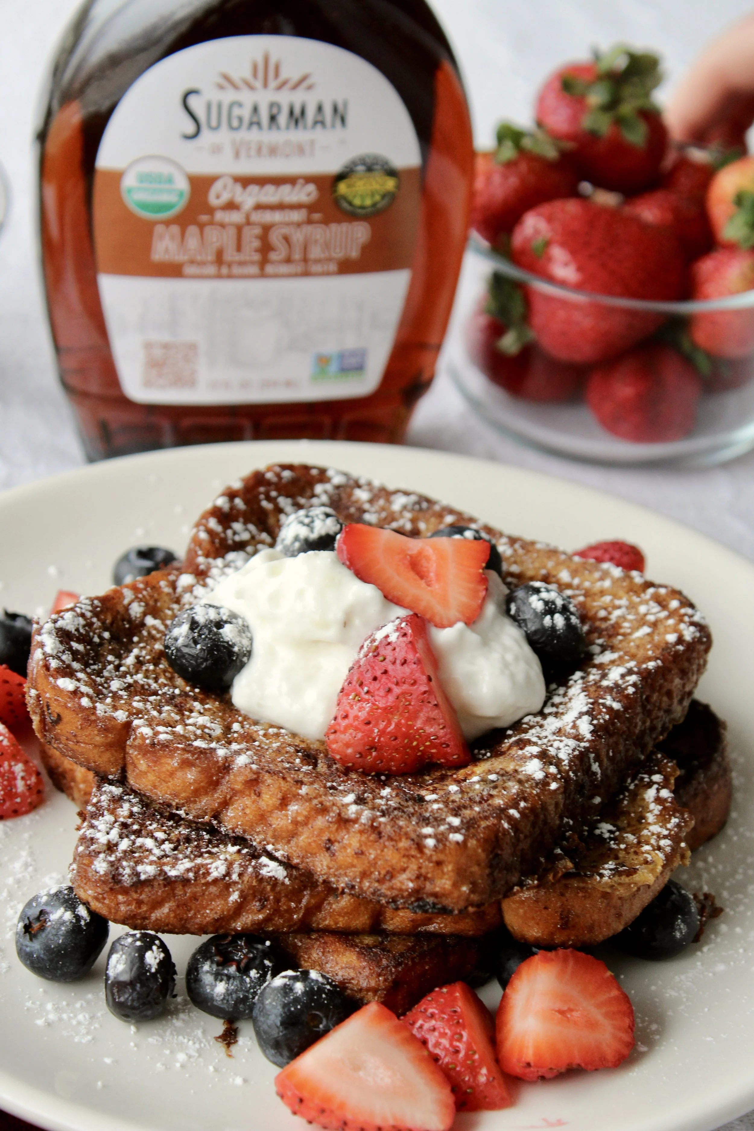 French toast topped with strawberries, blueberries, and whipped cream, served on a white plate with additional strawberries and blueberries, with a bottle of maple syrup and a bowl of strawberries in the background.