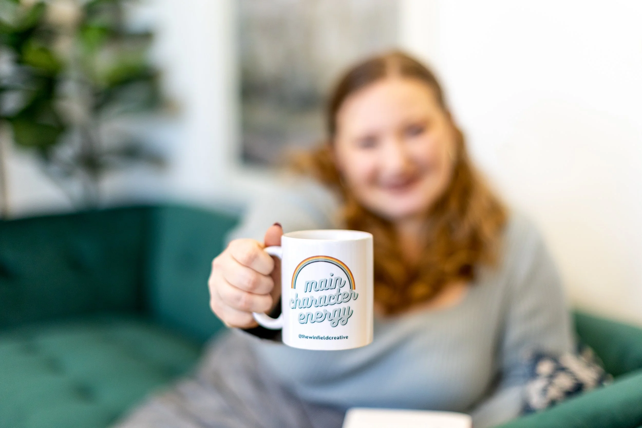 A woman with red hair smiling and holding a white coffee mug with the words "main character energy" and a rainbow graphic, in a cozy indoor setting.