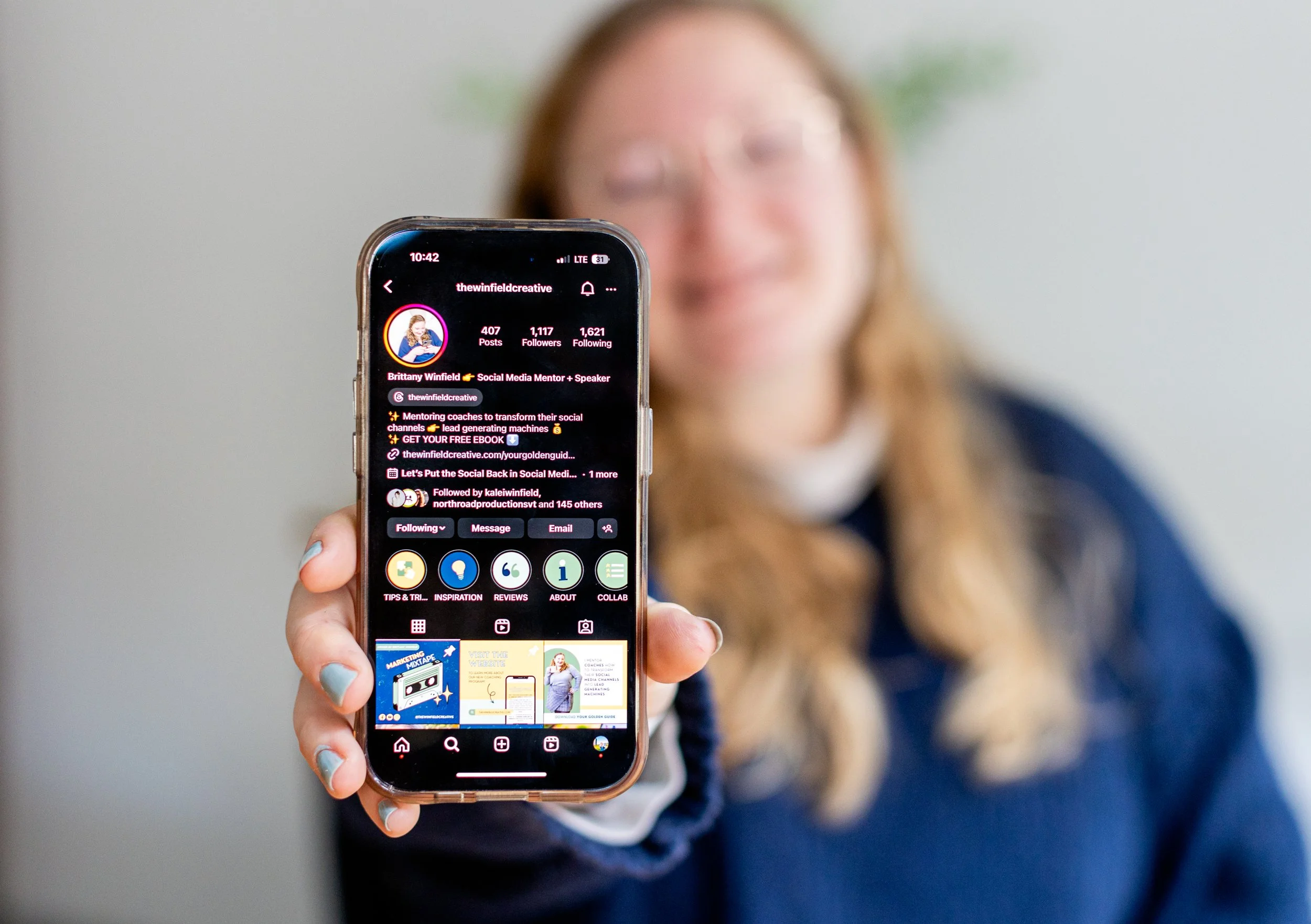 A woman with long blonde hair and glasses holding a smartphone with an Instagram profile page visible. The woman is smiling and blurred in the background, and the profile appears to belong to Brittanny Winfield, a social media mentor and speaker.