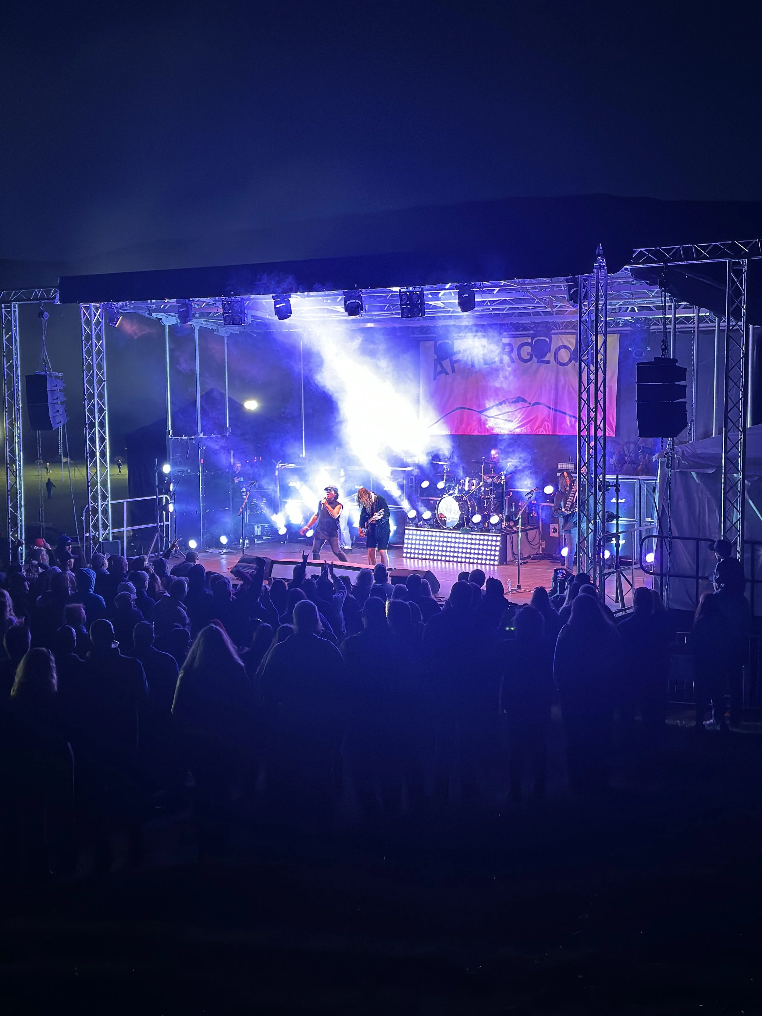 A live outdoor concert at night with a stage lit up in purple and white lights, a band performing, and a crowd watching.