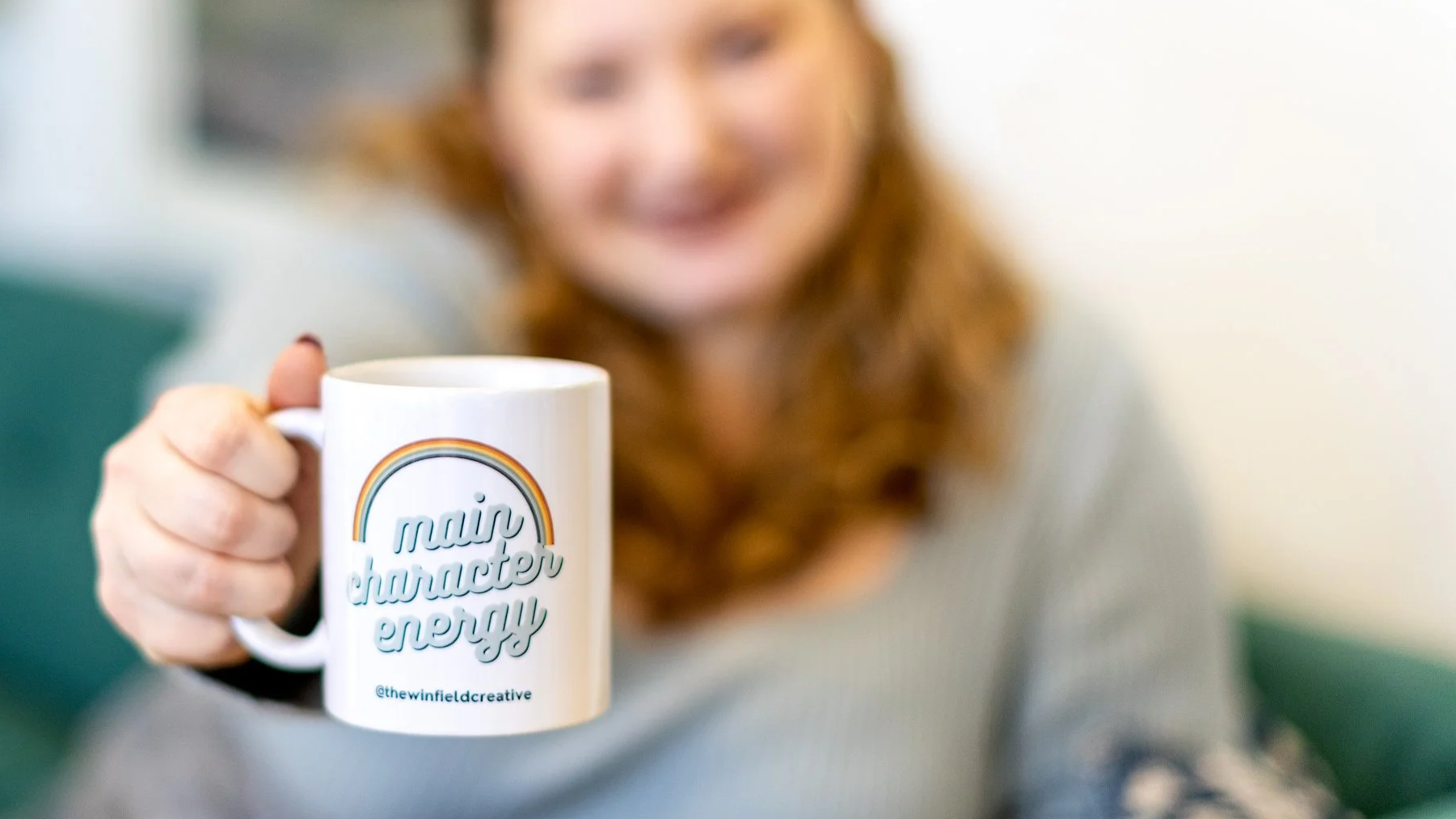 A woman with curly red hair smiling and holding a white mug with a rainbow design that says 'main character energy' in front of her face.