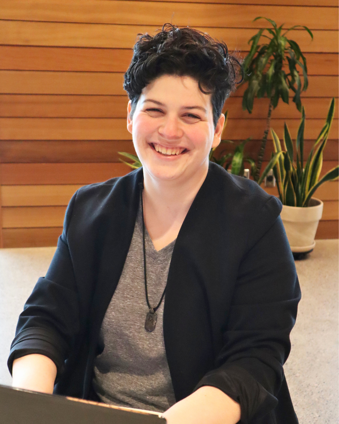 A smiling person with short, dark, curly hair sitting at a table in front of a wooden wall and potted plants.