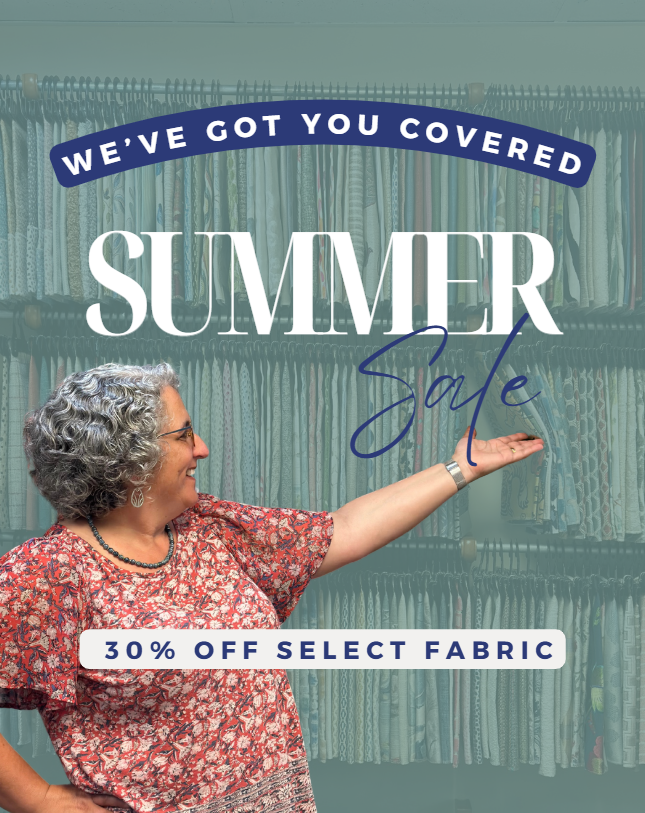 A woman with curly gray hair wearing glasses and a red patterned blouse points to a sign that says 'We've Got You Covered Summer Sale' and '30% Off Select Fabric' in a fabric store.