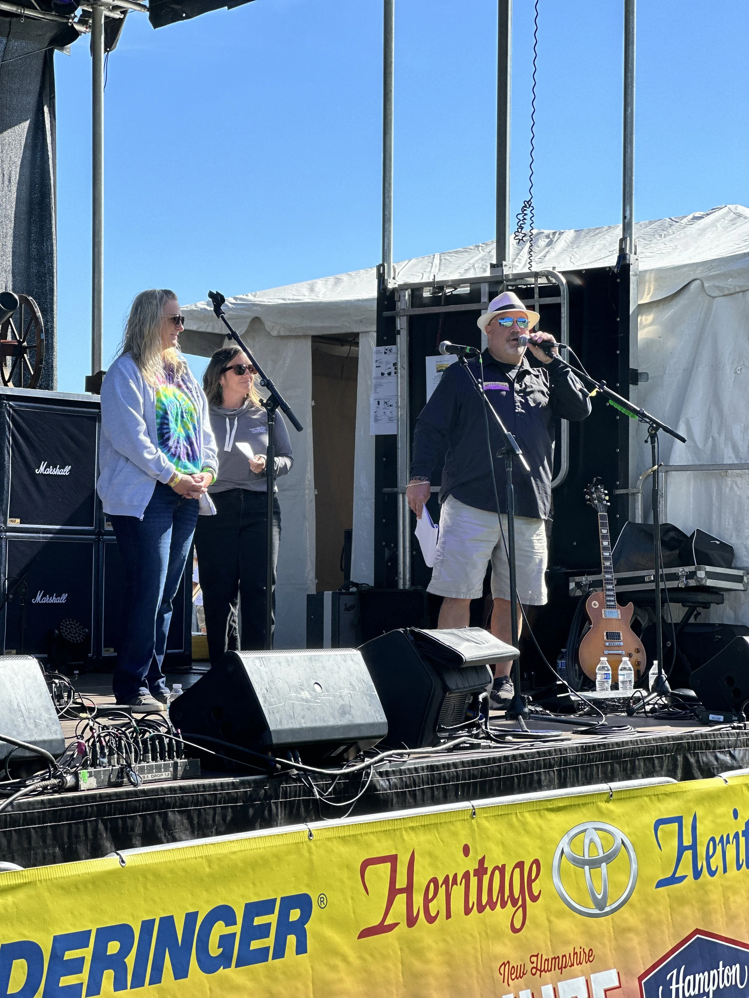 Three people on a stage outdoors with a man speaking into a microphone, two women standing beside him, and musical equipment including an electric guitar nearby. An event banner is visible at the front of the stage.