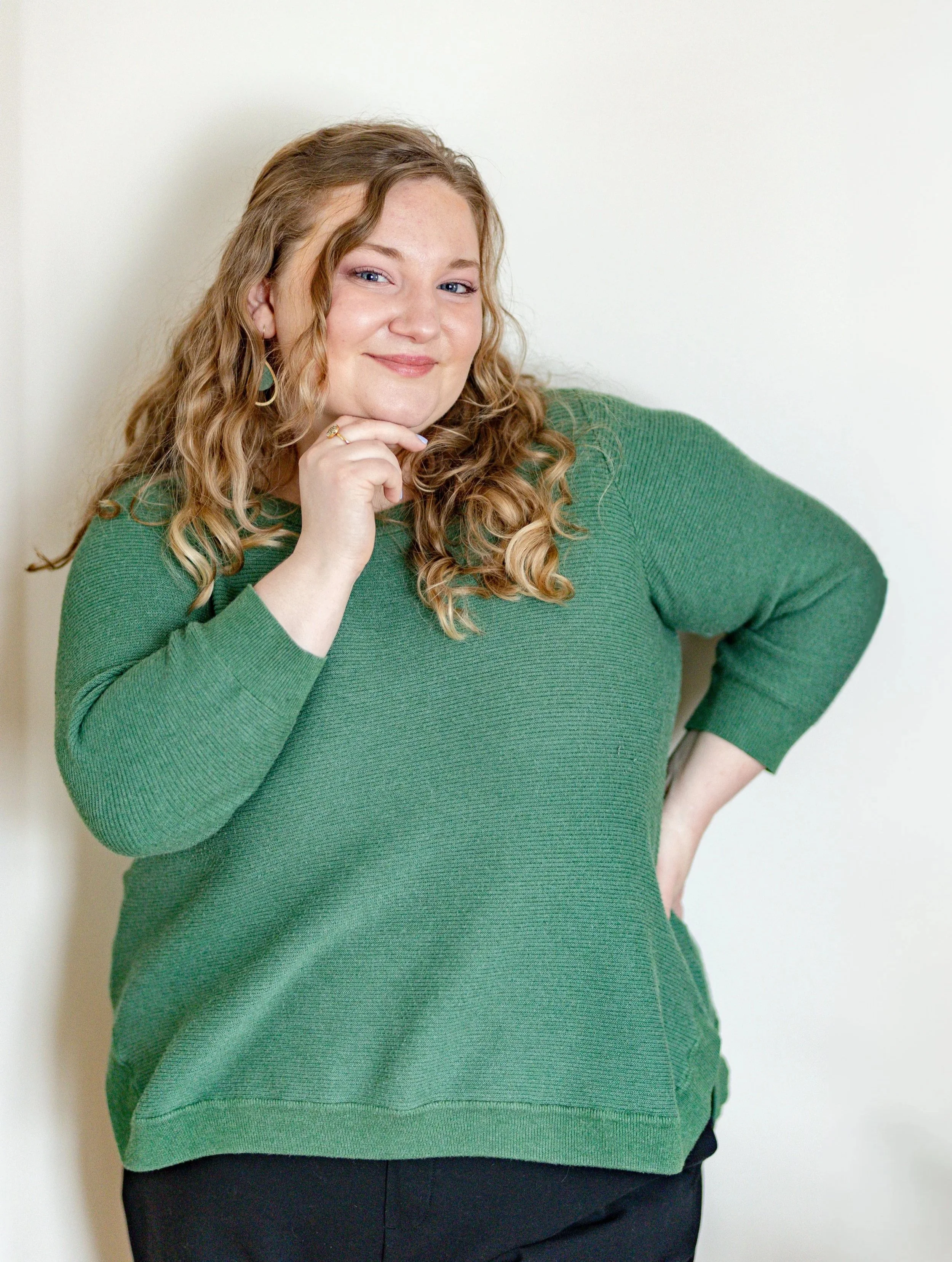 A woman with curly red hair, wearing a green sweater, smiling with her hand resting on her chin, standing against a plain white wall.
