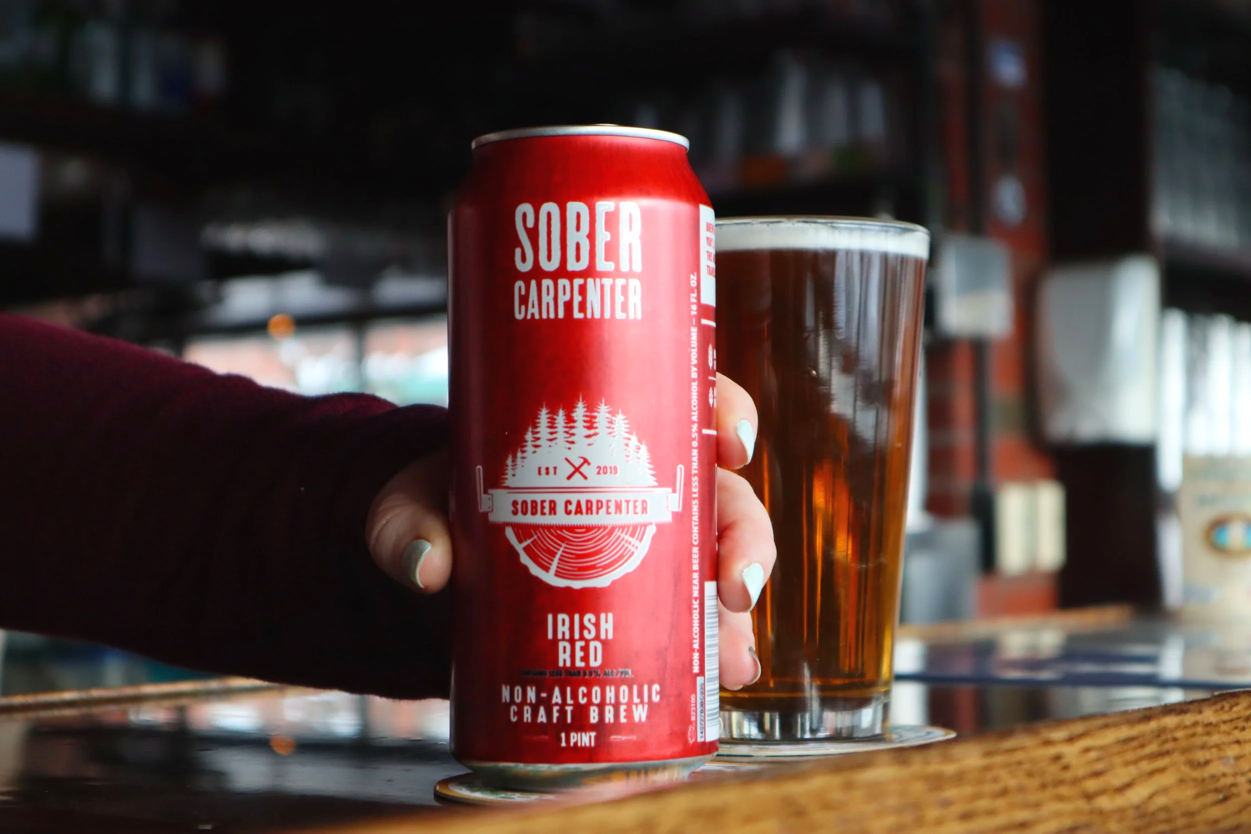 A person holding a red can of Sober Carpenter Irish Red non-alcoholic craft brew in front of a glass of beer on a wooden bar counter, with a background of a bar or brewery setting.