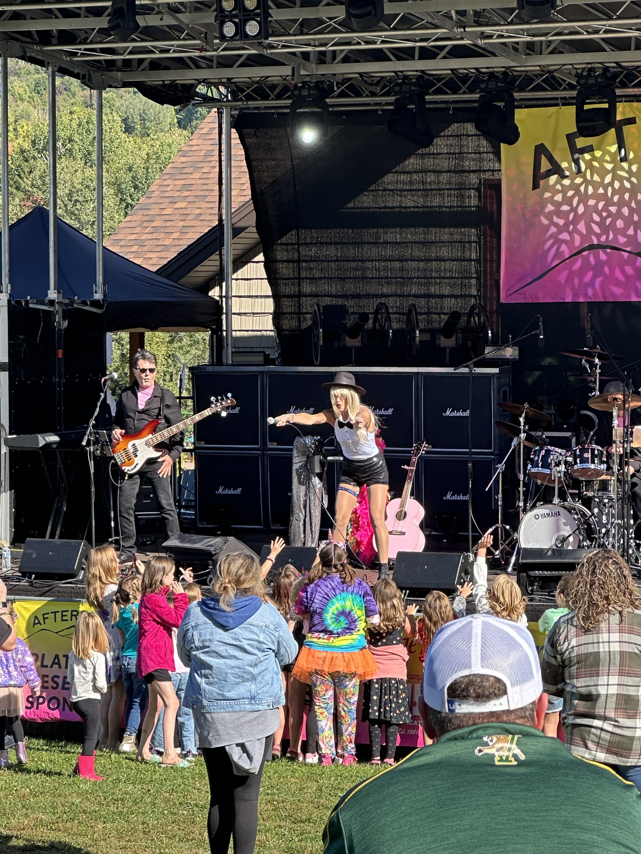 A live outdoor music performance with a band on stage and children dancing in front. The stage has guitars, drums, and amplifiers. The background shows a building roof and green trees.