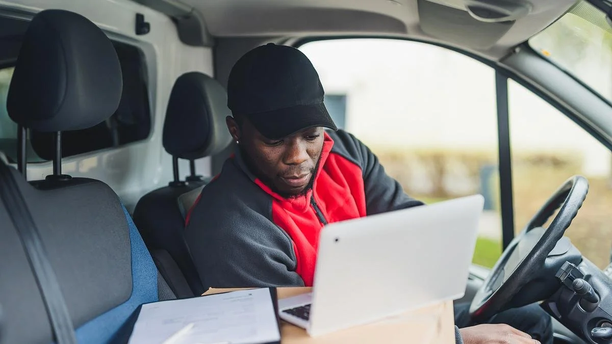 A man in a black cap and red jacket working on a laptop inside a vehicle.