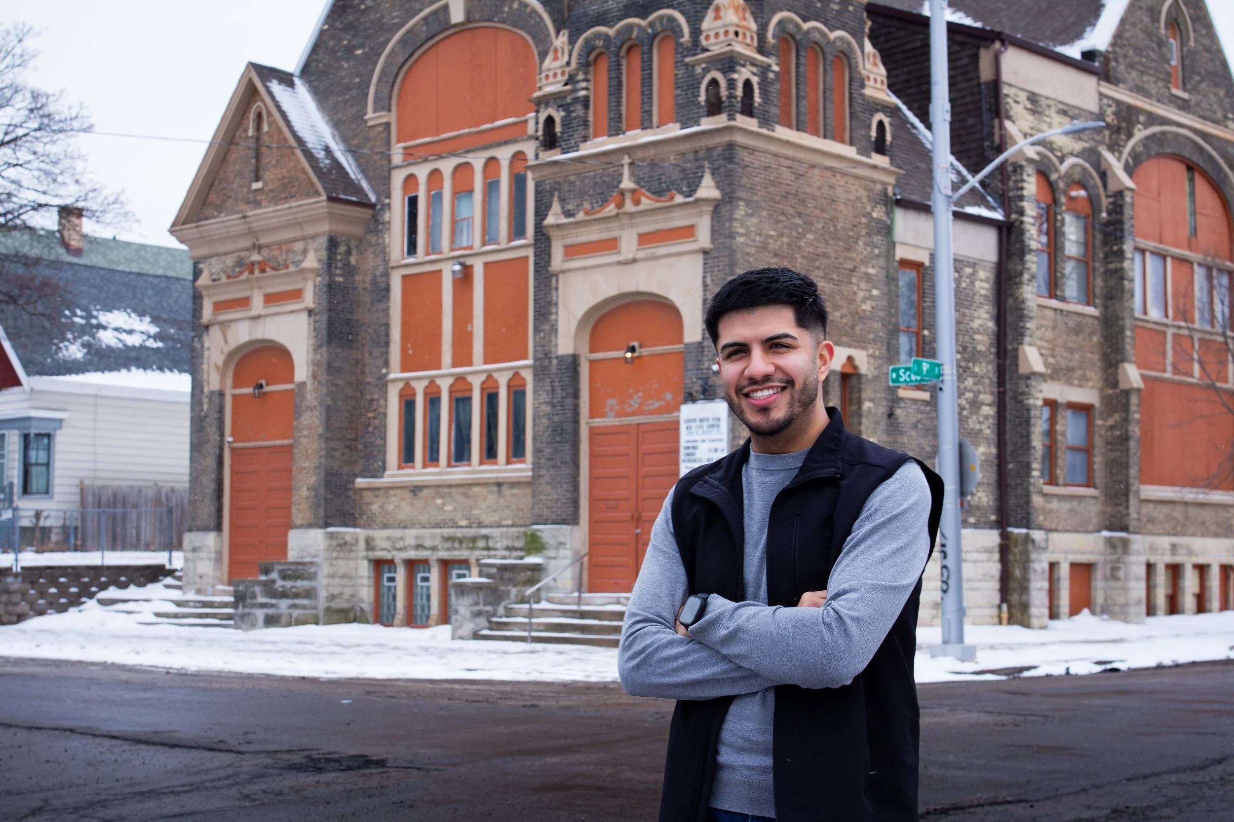 A young man with dark hair and a beard (Ismael Luna) smiling with arms crossed, wearing a grey shirt and black jacket, standing on a snowy street in front of an old brick building with orange panels and large arched windows.