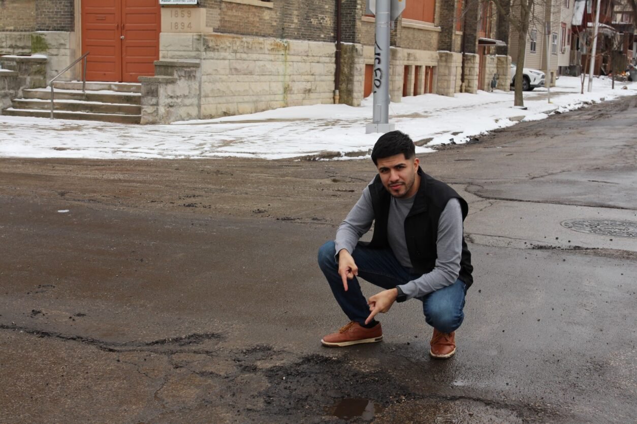 A man crouching on a city street with potholes, snow on the sidewalk, and buildings in the background.