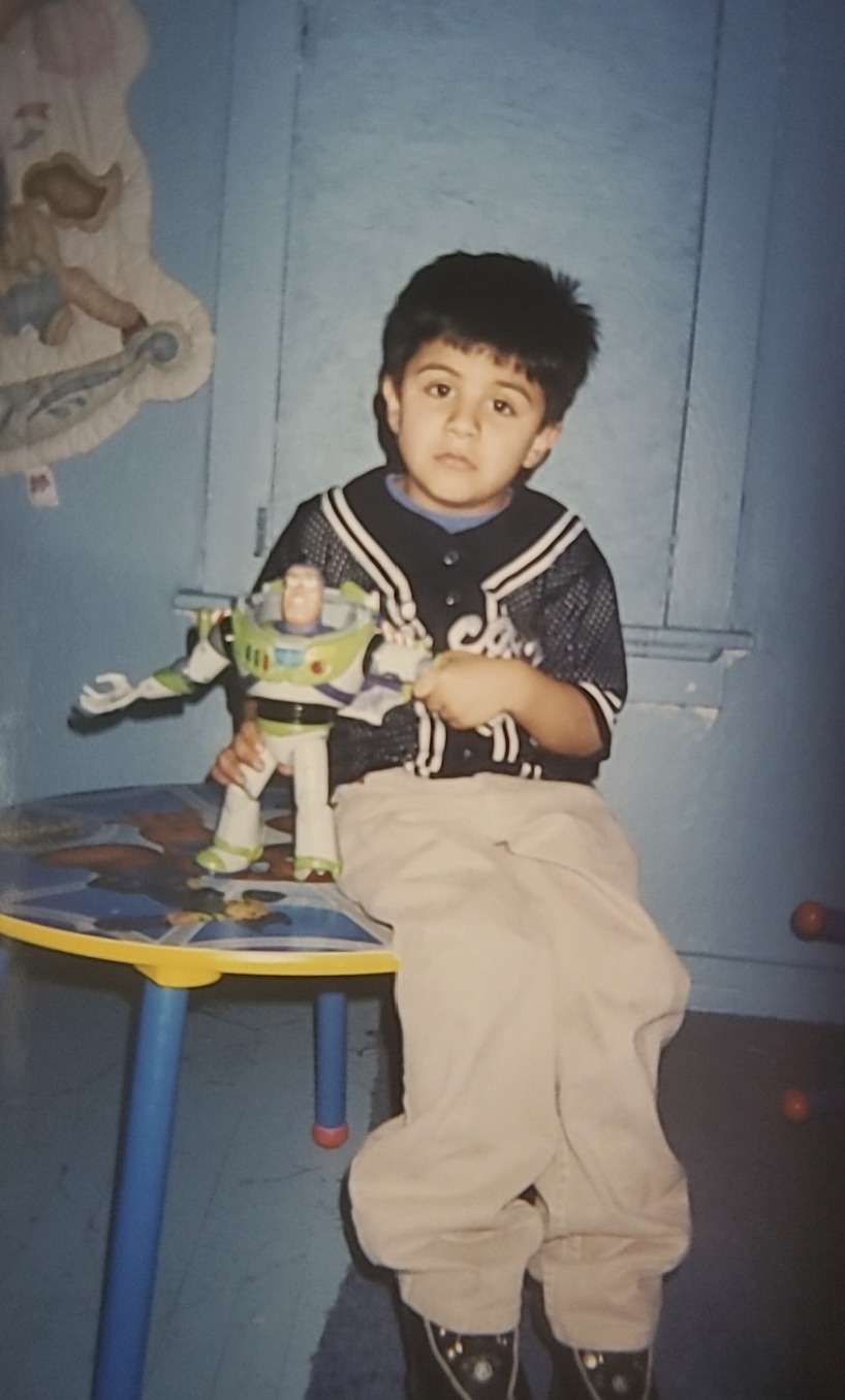 A young boy (Ismael Luna) with black hair sits on a small chair in a blue room, holding a Buzz Lightyear toy. There is a wall hanging on the left side of the image.