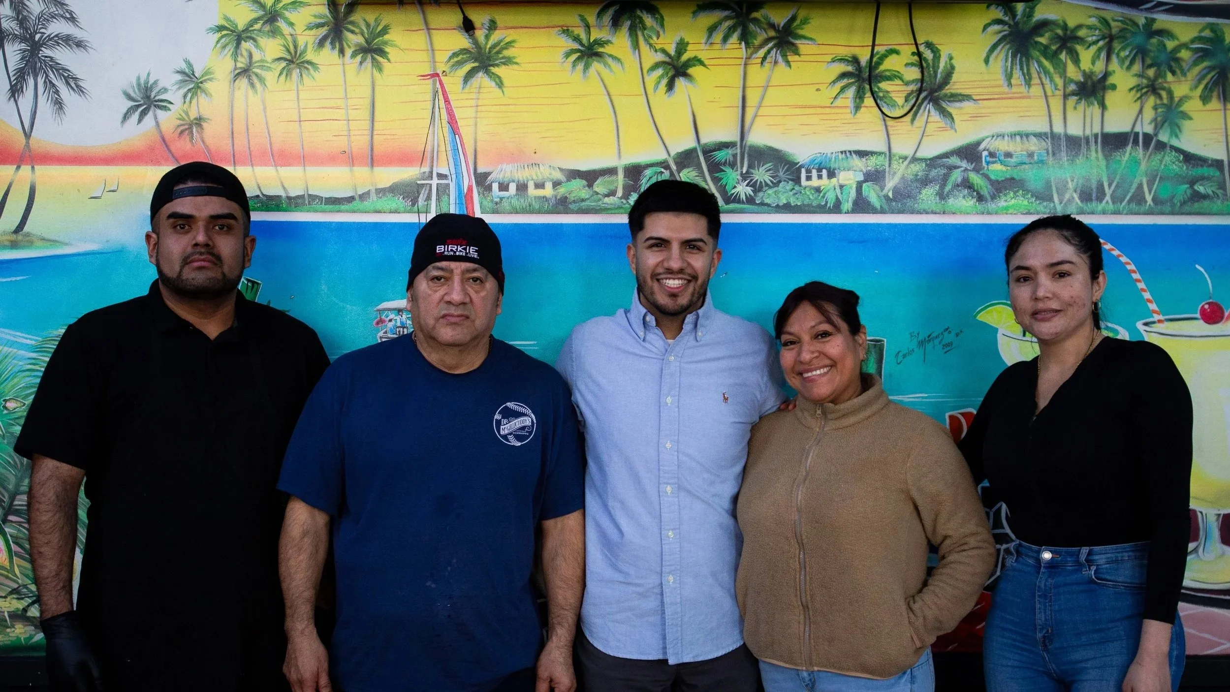 Five people standing in front of a tropical mural with palm trees, a boat, and drinks, smiling at the camera.